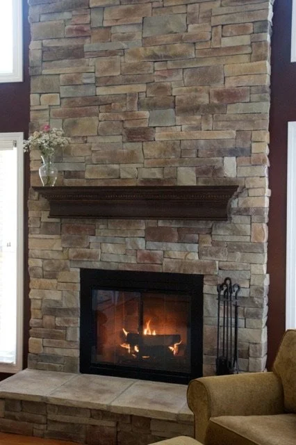 A stone fireplace with a wooden mantel, a fire burning inside the hearth, and a vase with flowers on top of the mantel.