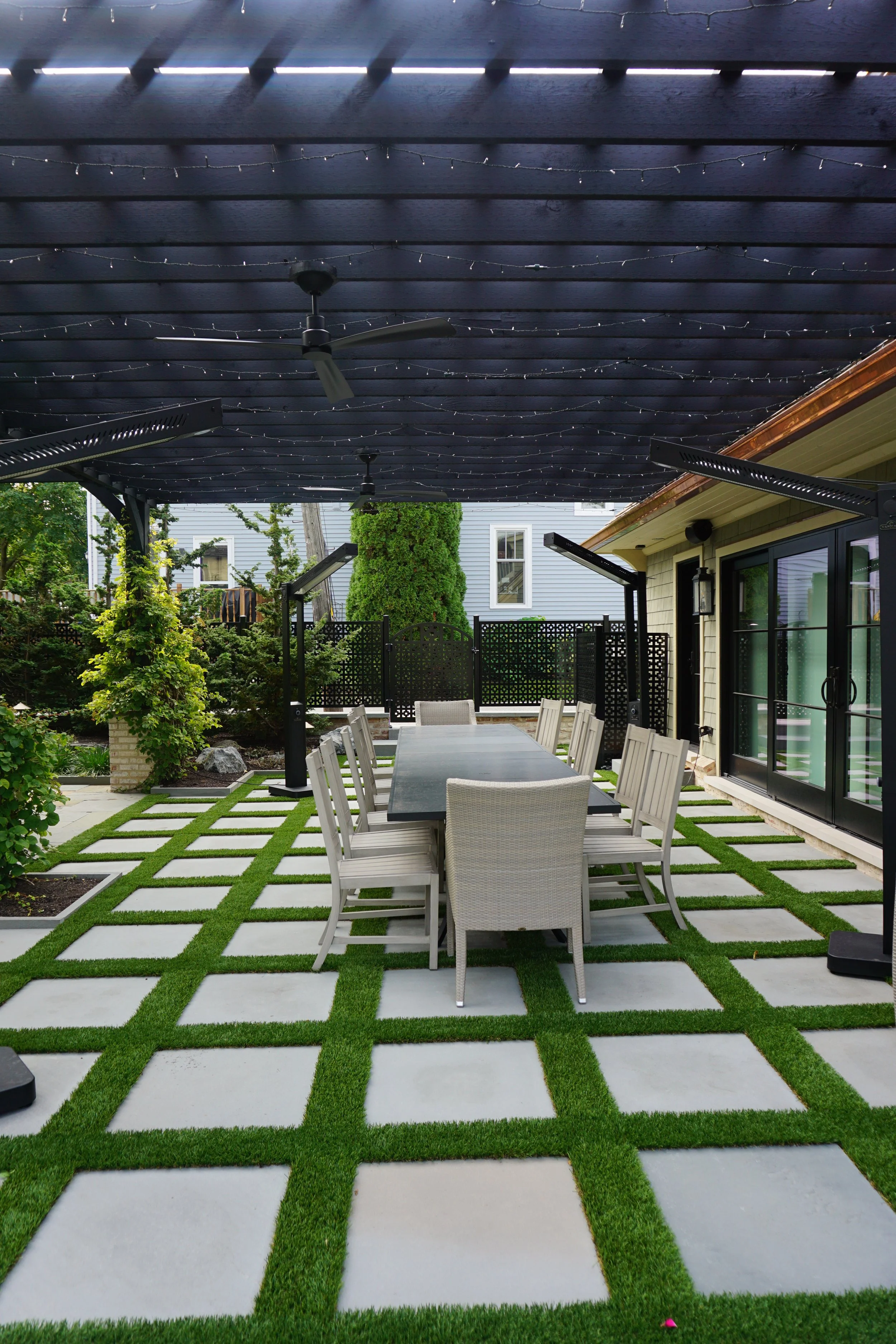 Backyard patio with outdoor dining table and chairs under a dark wooden pergola decorated with string lights and ceiling fans. The ground has a checkerboard pattern of grass and white concrete tiles. Green plants and trees are visible in the background, along with a black privacy fence.