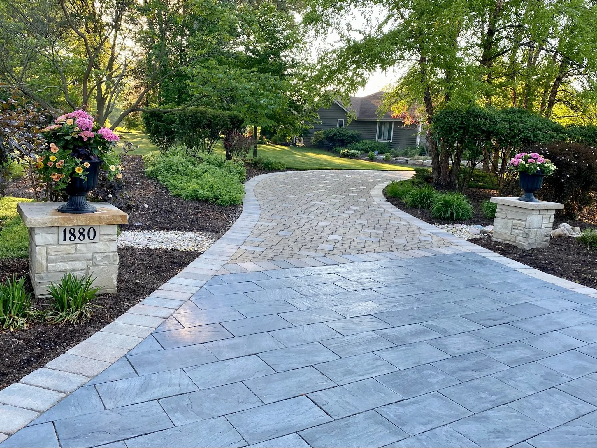 A curved brick pathway leading to a house, flanked by flower pots with pink and yellow flowers, green bushes, and trees in a well-maintained garden on a sunny day.