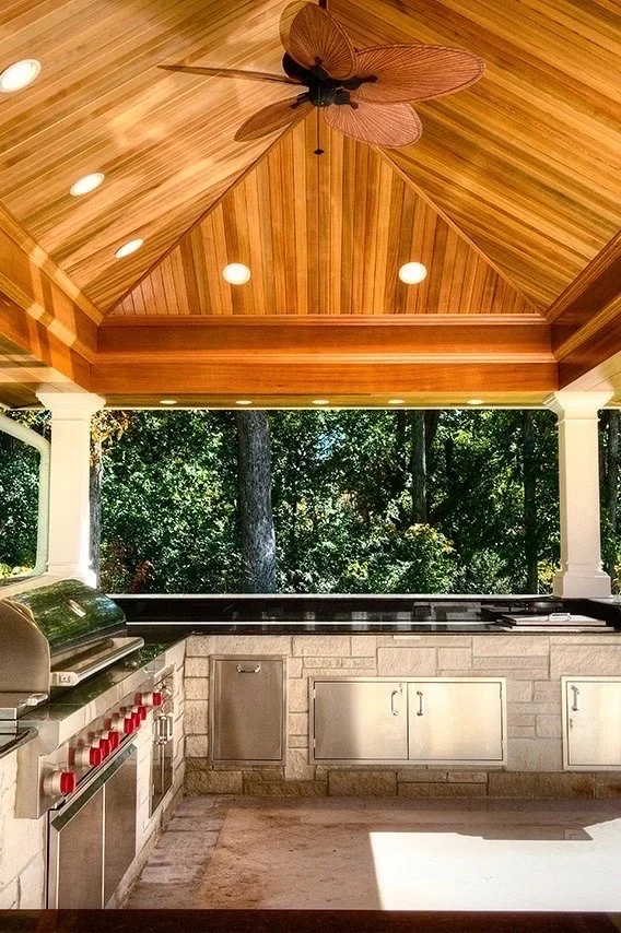 Outdoor kitchen with a wood-paneled ceiling, ceiling fan shaped like a butterfly, stainless steel appliances, stone counter, and a view of green trees outside.