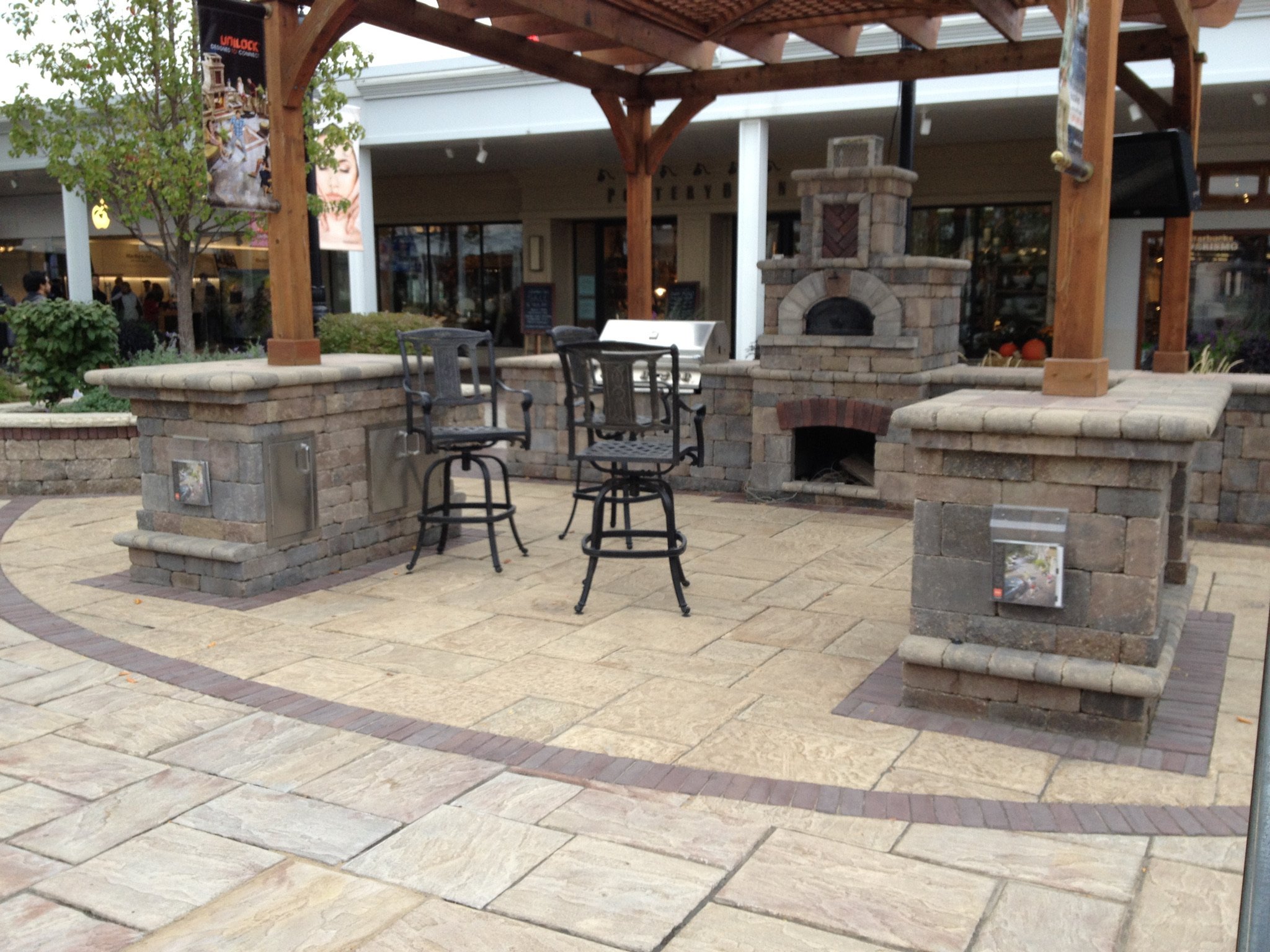 An outdoor brick and stone patio area with a wooden pergola. There are metal chairs and a metal barbecue grill. A brick oven or fireplace is built into the brick wall. In the background, shop windows and people walking by can be seen.