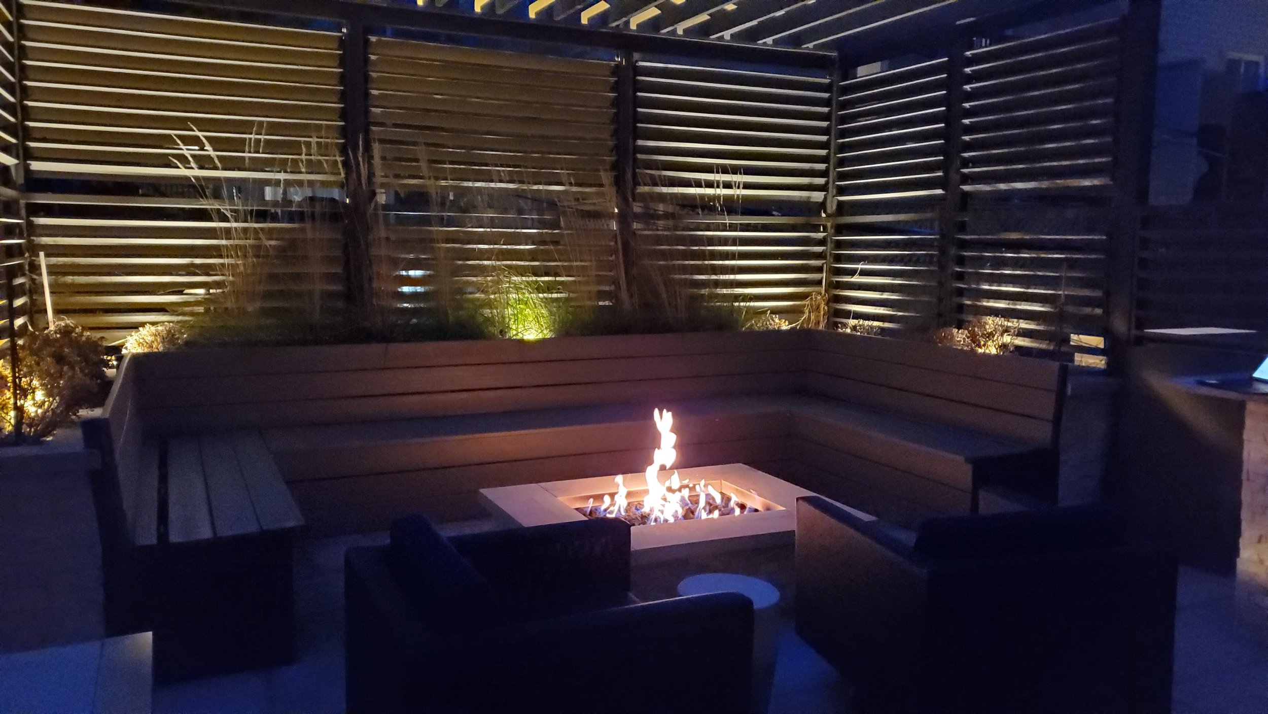 Nighttime view of a backyard patio with a fire pit, wooden benches, and a privacy fence with horizontal slats, illuminated by outdoor lighting.