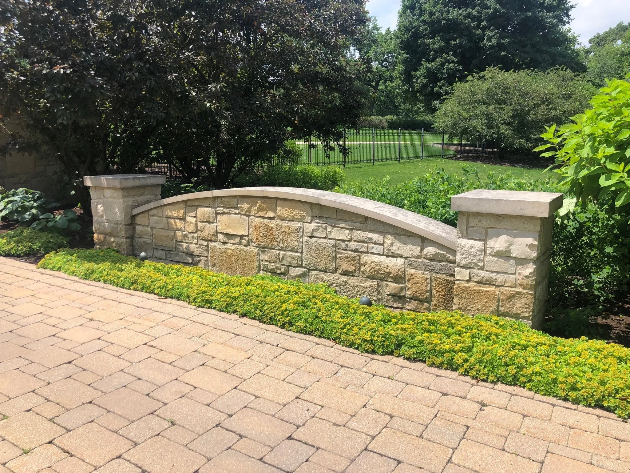 A stone and brick low wall with a curved top edge, separating a paved walkway from a lush garden with various green plants. Behind the wall, there are tall leafy trees, shrubs, and a black metal fence. The sky is mostly clear with some clouds.