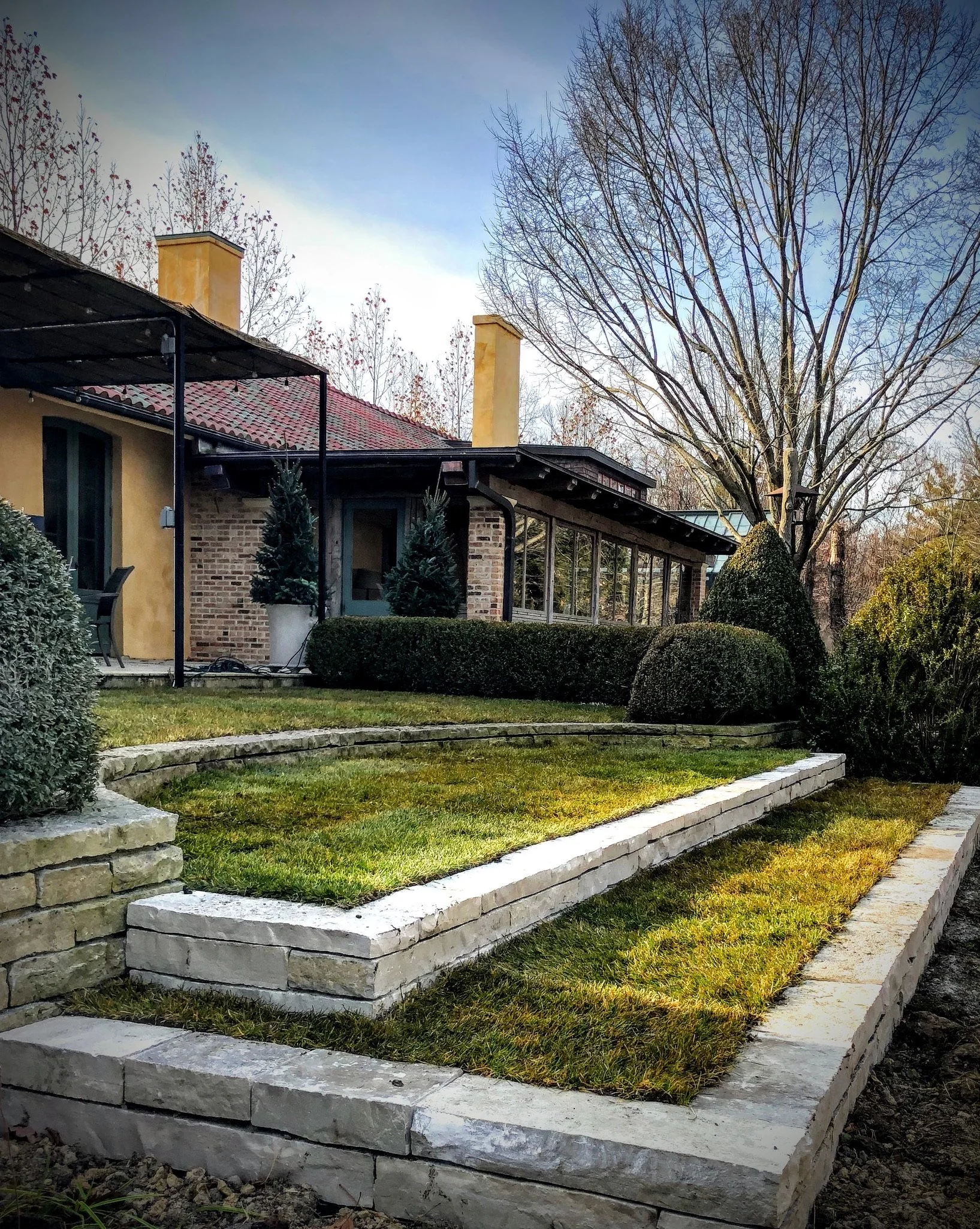 A landscaped yard with a tiered stone retaining wall, grass, and a pathway, with a house featuring a brick and stucco exterior, a red-tiled roof, and a covered porch. There are trees, some with no leaves, and shrubs around the yard.