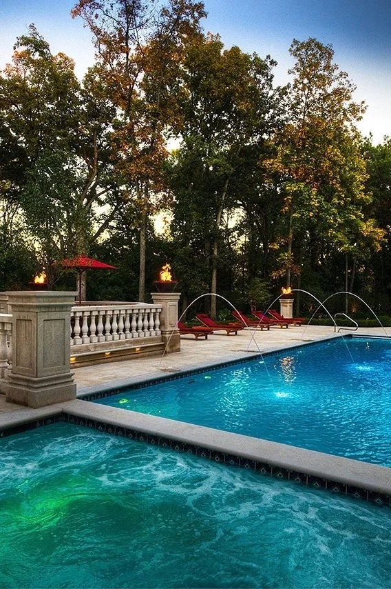 A backyard swimming pool with a hot tub in the foreground, surrounded by a stone deck, red lounge chairs, and trees in the background during sunset.