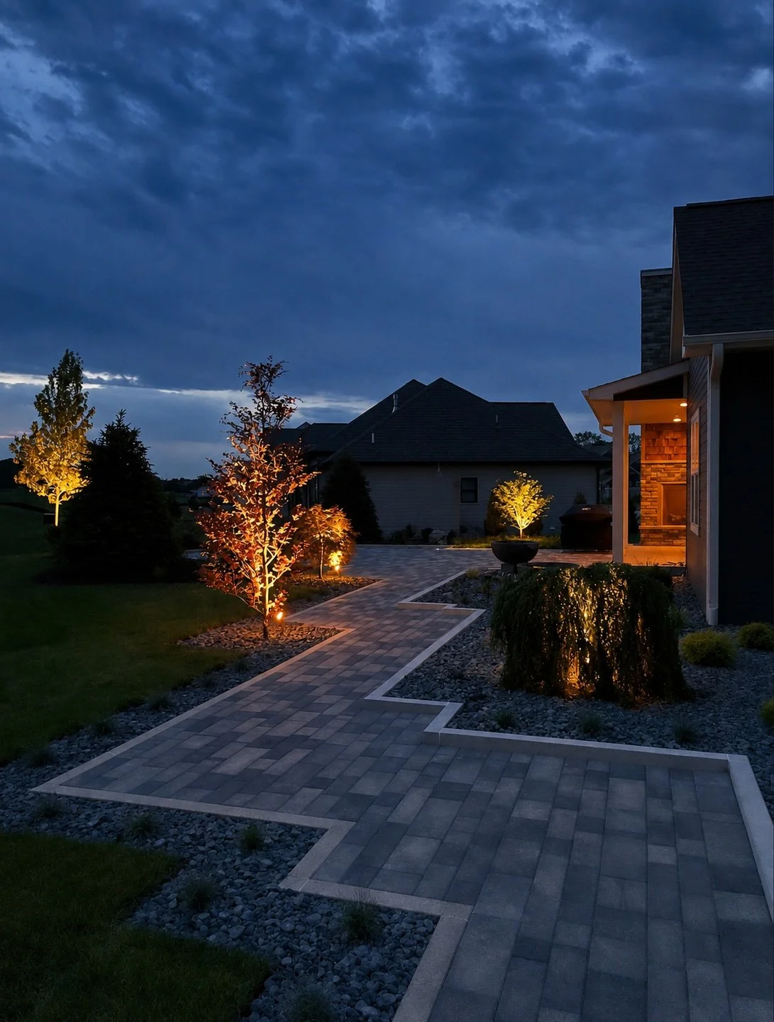 Nighttime view of a landscaped backyard with illuminated trees and a paved walkway leading to a house with outdoor lighting under a cloudy sky.