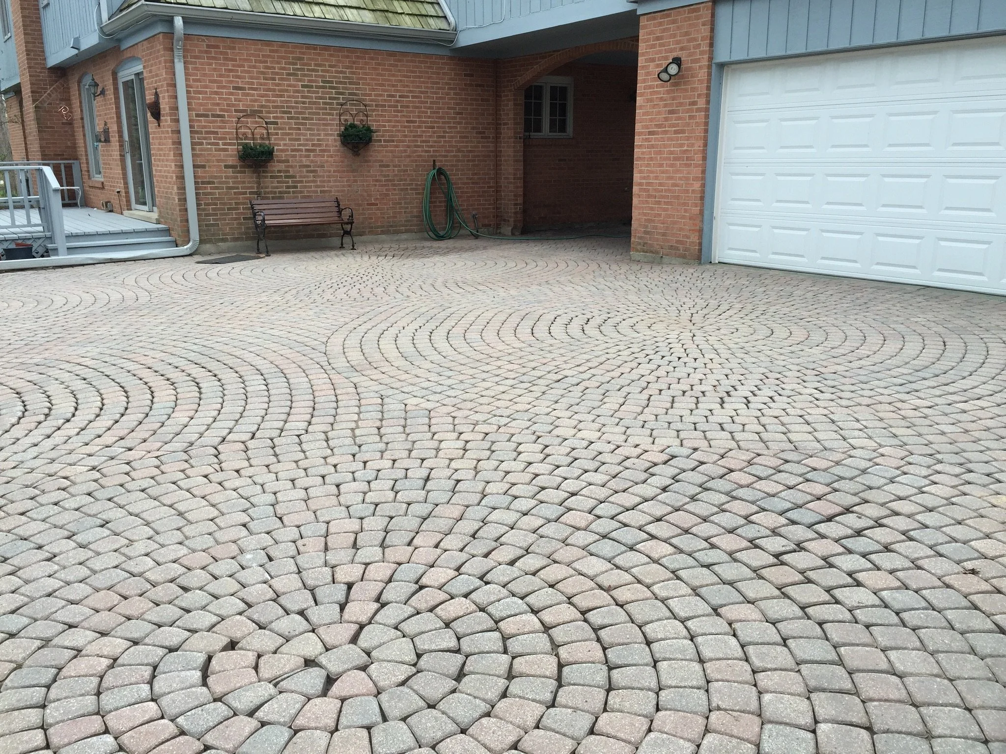 View of a house with a red brick wall, a white garage door, a small porch with stairs, a garden hose hanging on the wall, a wooden bench, and decorative wall planters, with a paved driveway in the foreground.
