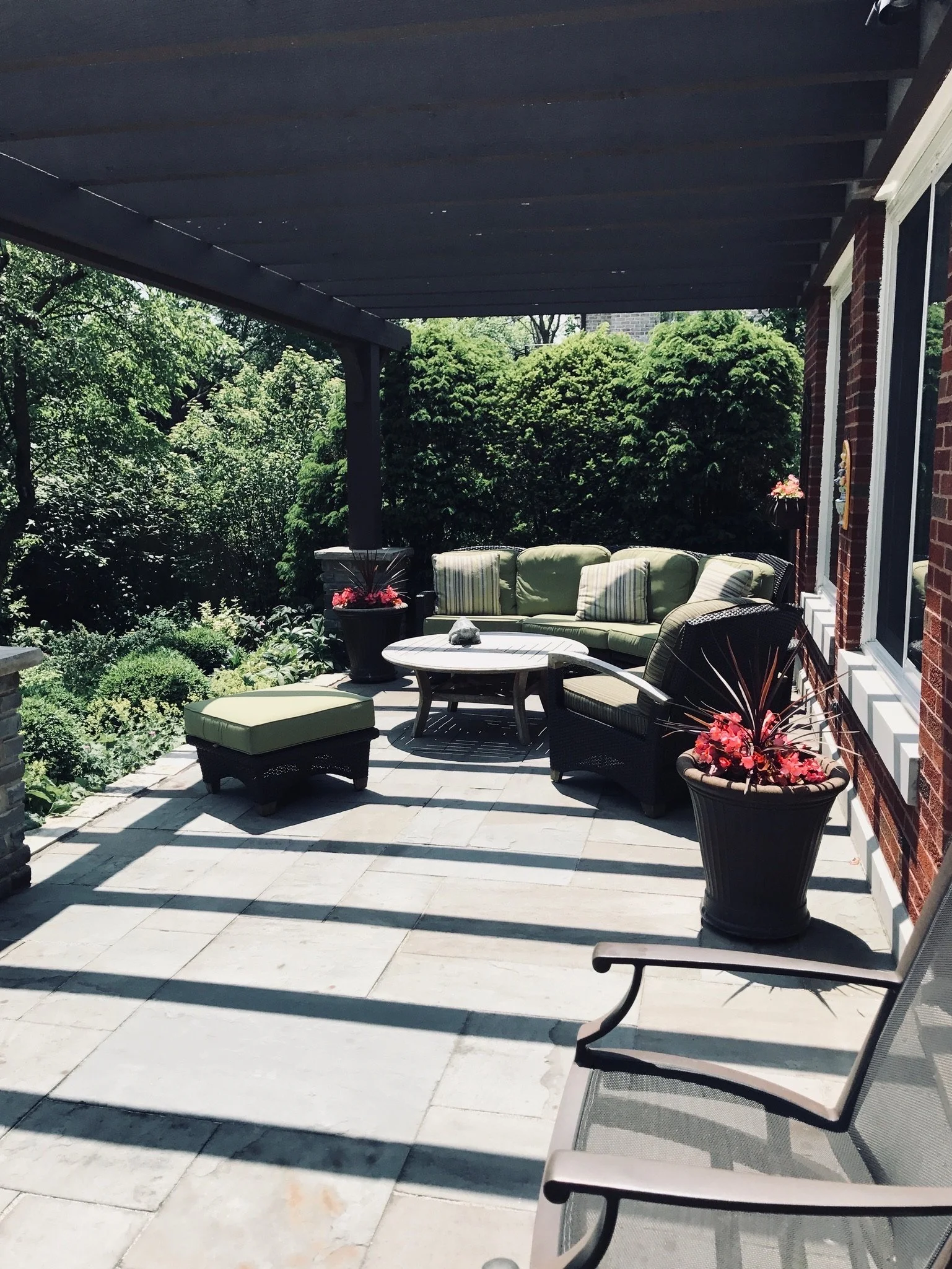 Back porch area with outdoor furniture including cushioned chairs and a table, decorated with potted plants and surrounded by green trees and shrubs.