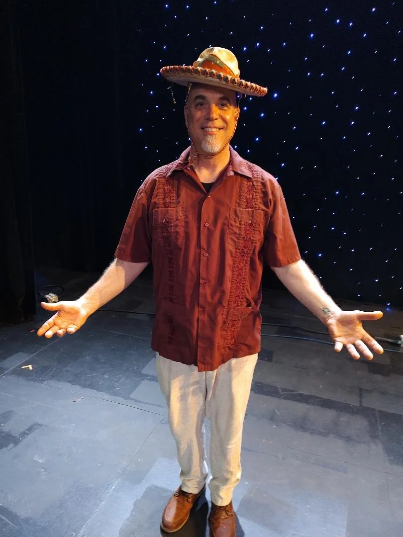 A man wearing a large sombrero hat, a maroon short-sleeved shirt, beige pants, and brown shoes, standing on a dark stage with black curtains and blue star-like lights in the background, smiling with arms open.