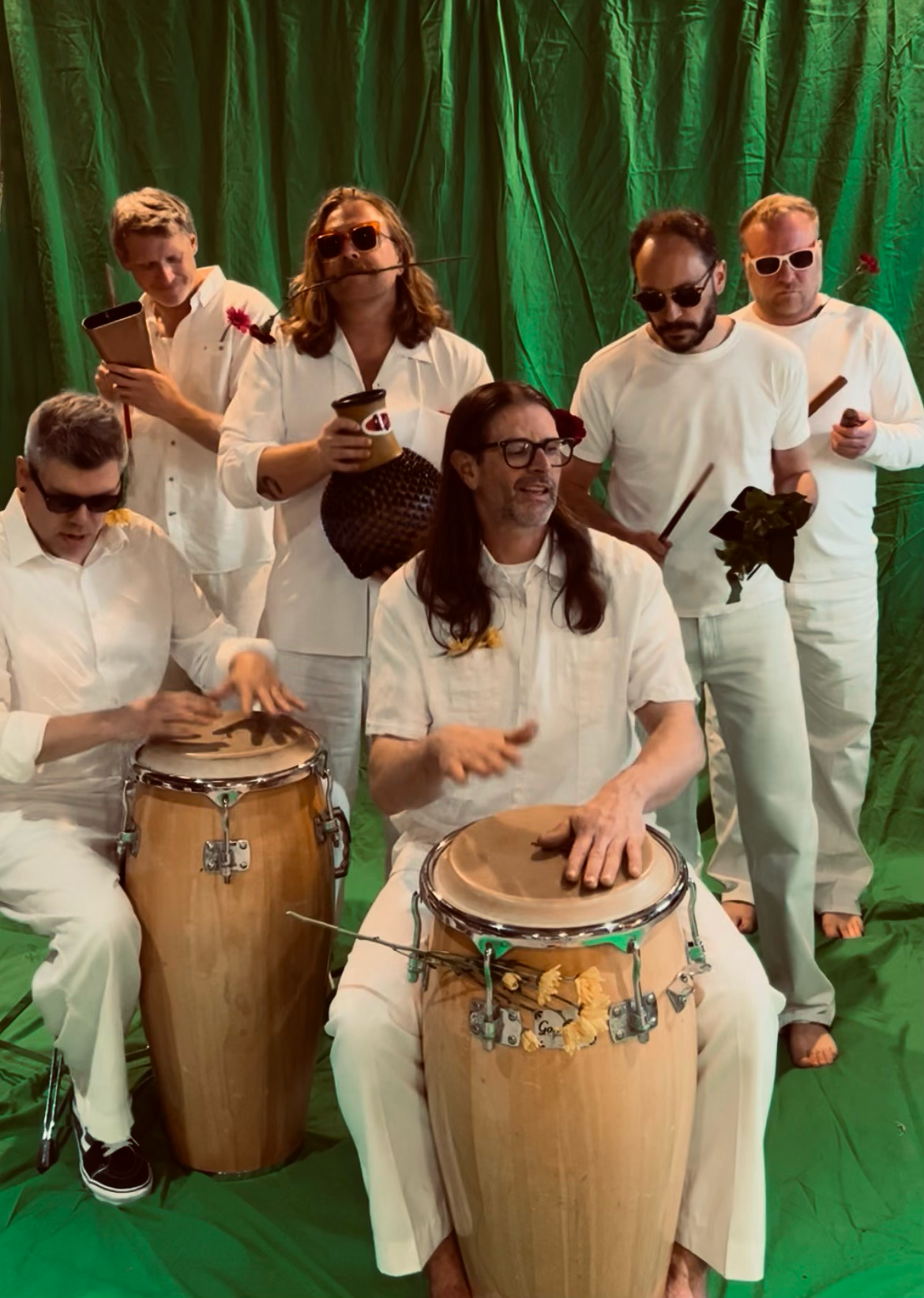 A group of seven men dressed in white shirts and white pants, some wearing sunglasses, gathered in front of a green curtain. Two men in the front are playing drums, while the others are holding various objects such as a pot, a leaf, and a stick. The scene appears lively and musical.