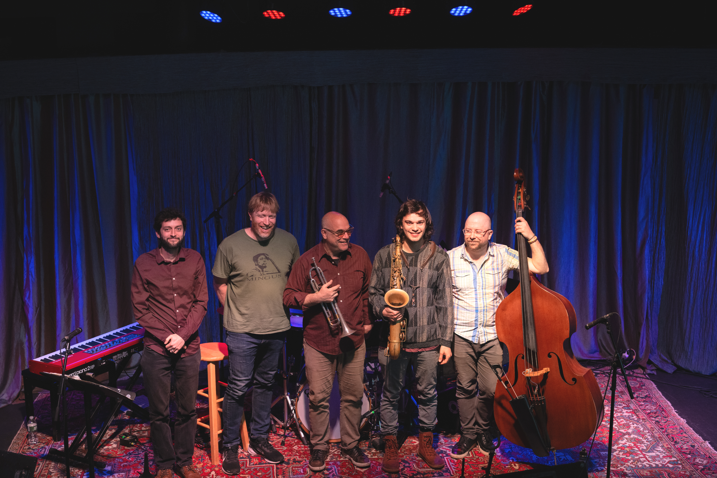 A group of five male musicians standing on a stage with dark blue curtains, holding musical instruments including a trumpet, saxophone, double bass, and standing near a keyboard. The stage is lit with red and blue stage lights.