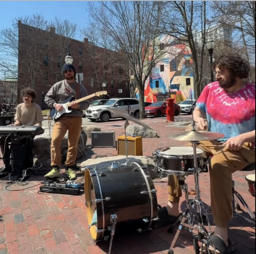 Three musicians performing outdoors on a sunny day, with cars parked behind them, a colorful mural on a building, leafless trees, and vintage-style street lamps in the background.