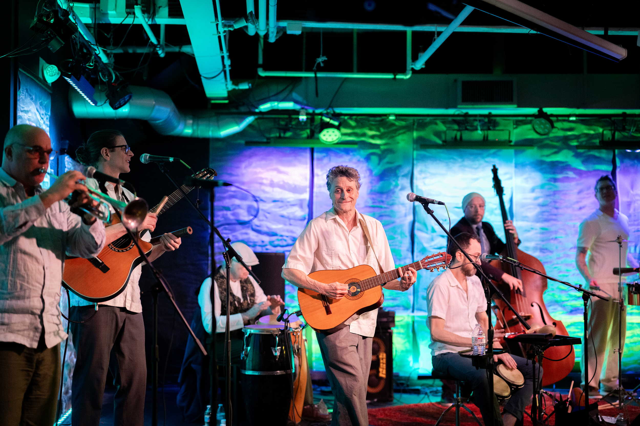 A group of musicians performing on stage, with some playing guitars and others playing various instruments, under colorful stage lighting.