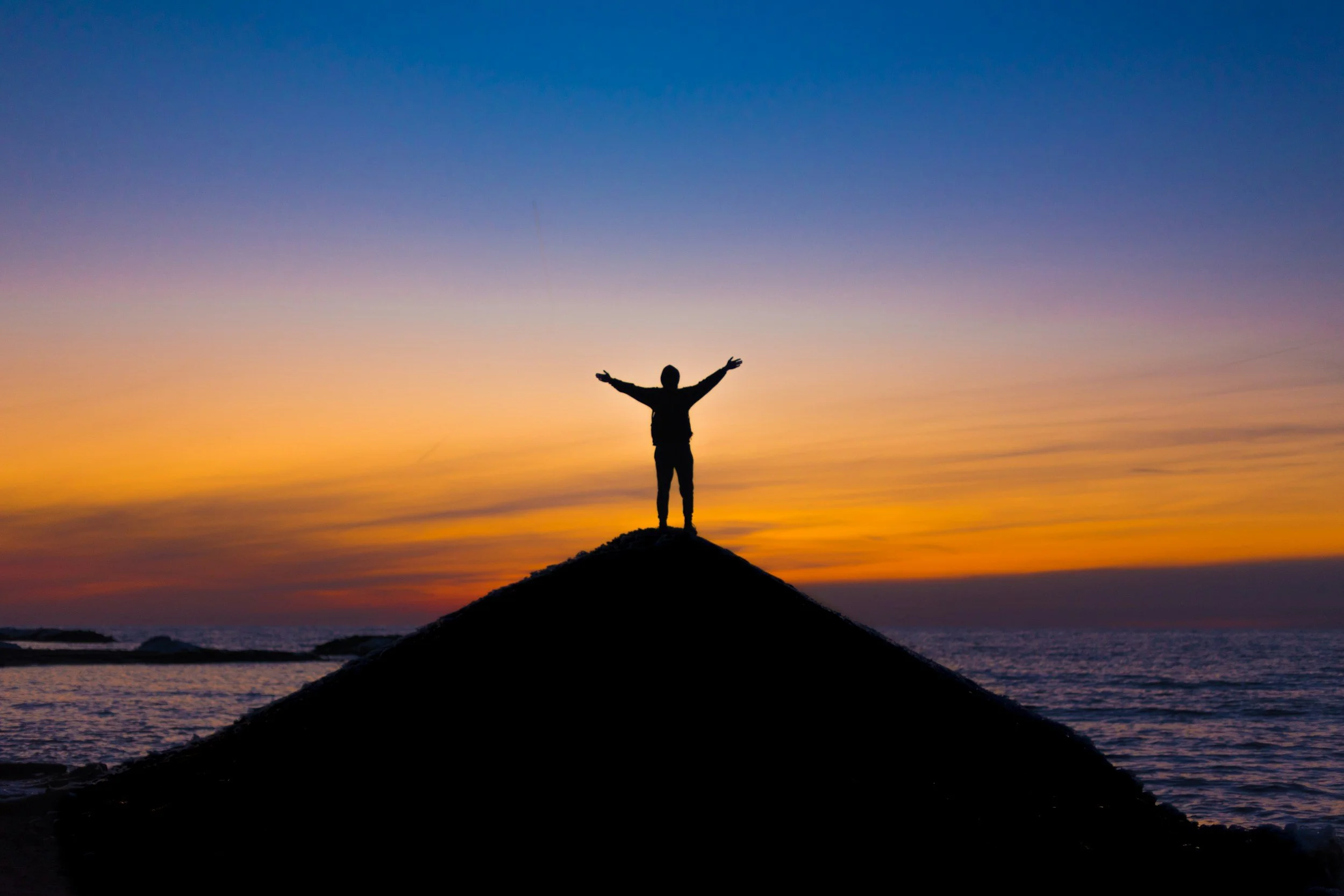 Person standing with arms outstretched on a hill at sunset with colorful sky and ocean in background.