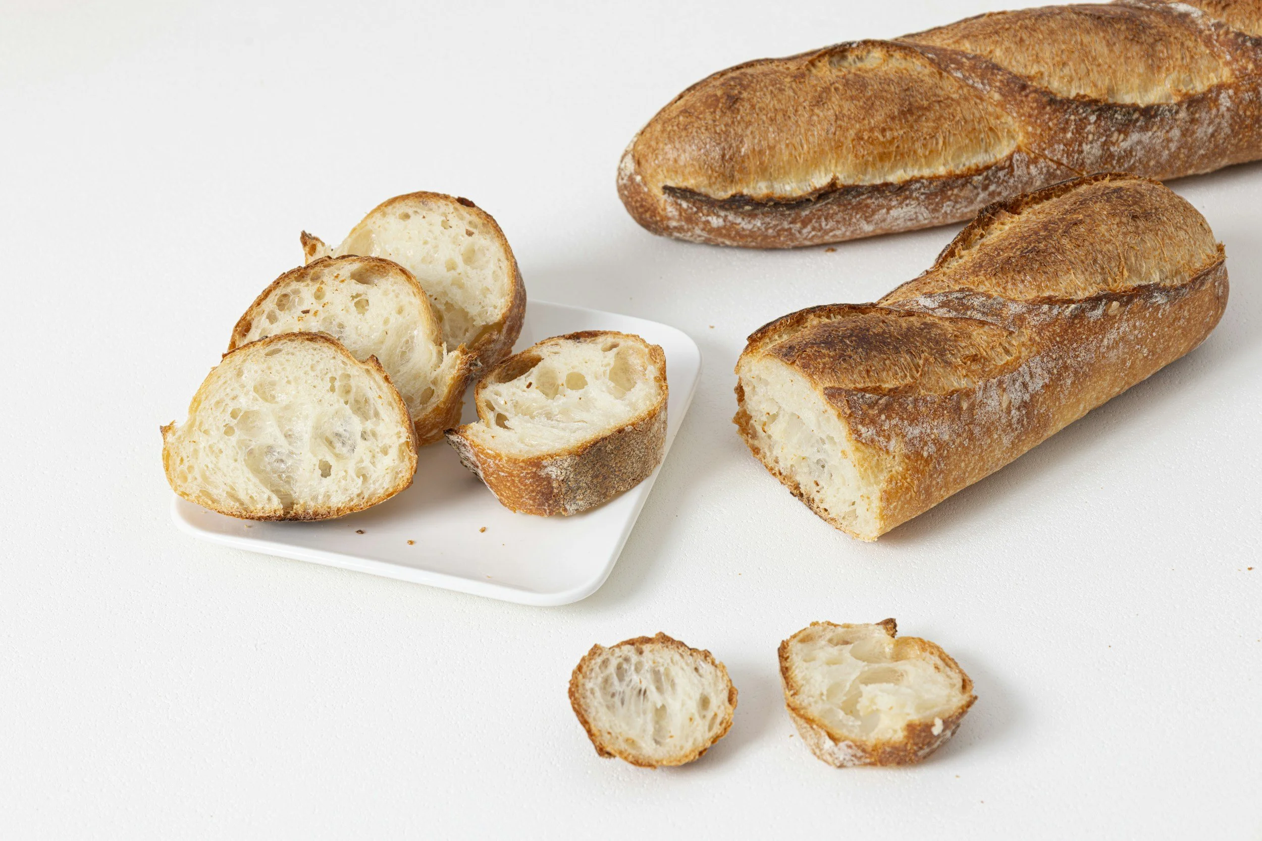 Three baguettes, two whole and one partly sliced, on a white surface with some slices on a white plate