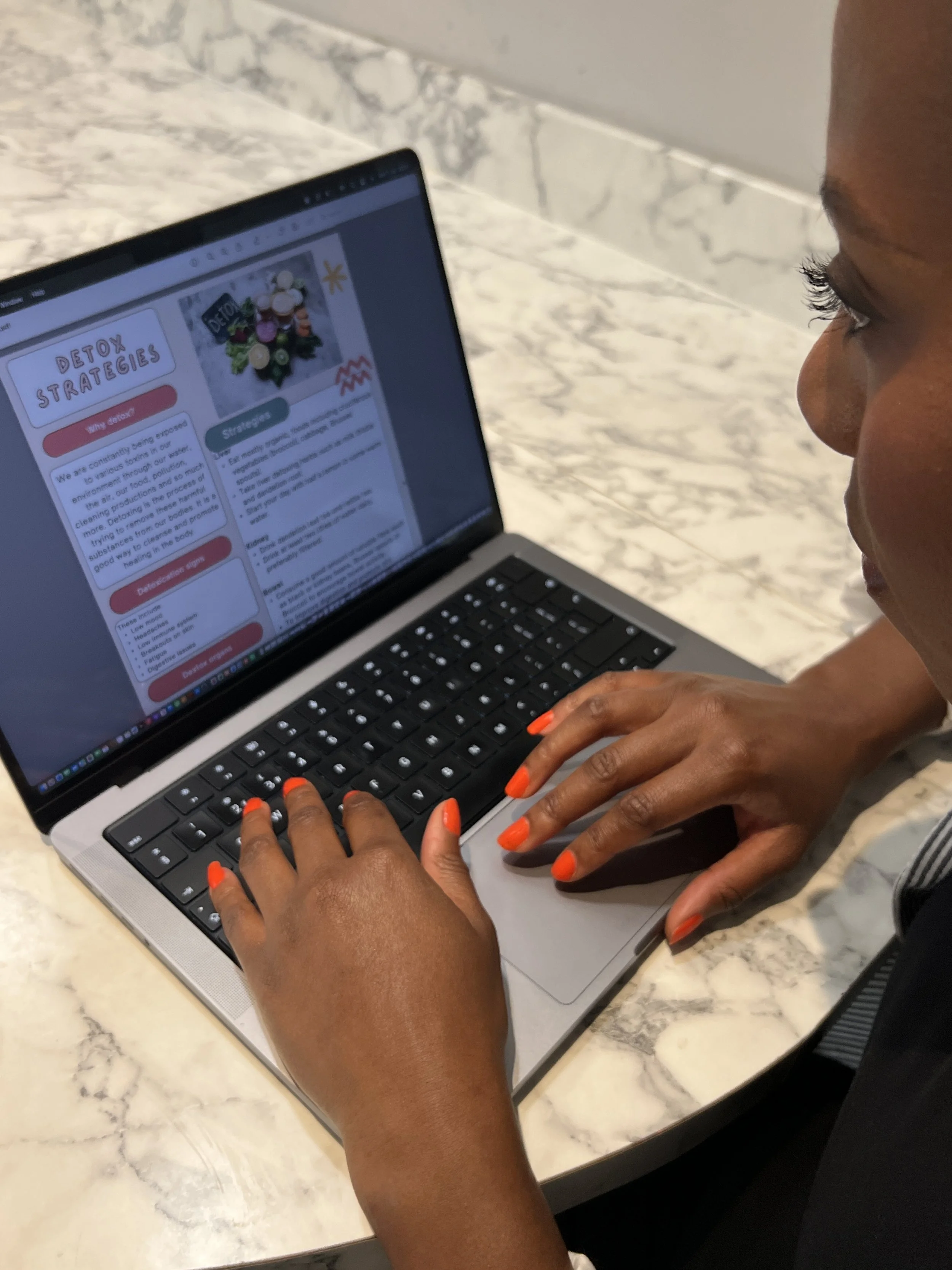A person with dark skin and bright orange nail polish is typing on a laptop with a marble countertop in the background. The screen displays an article titled 'Detox Strategies'.