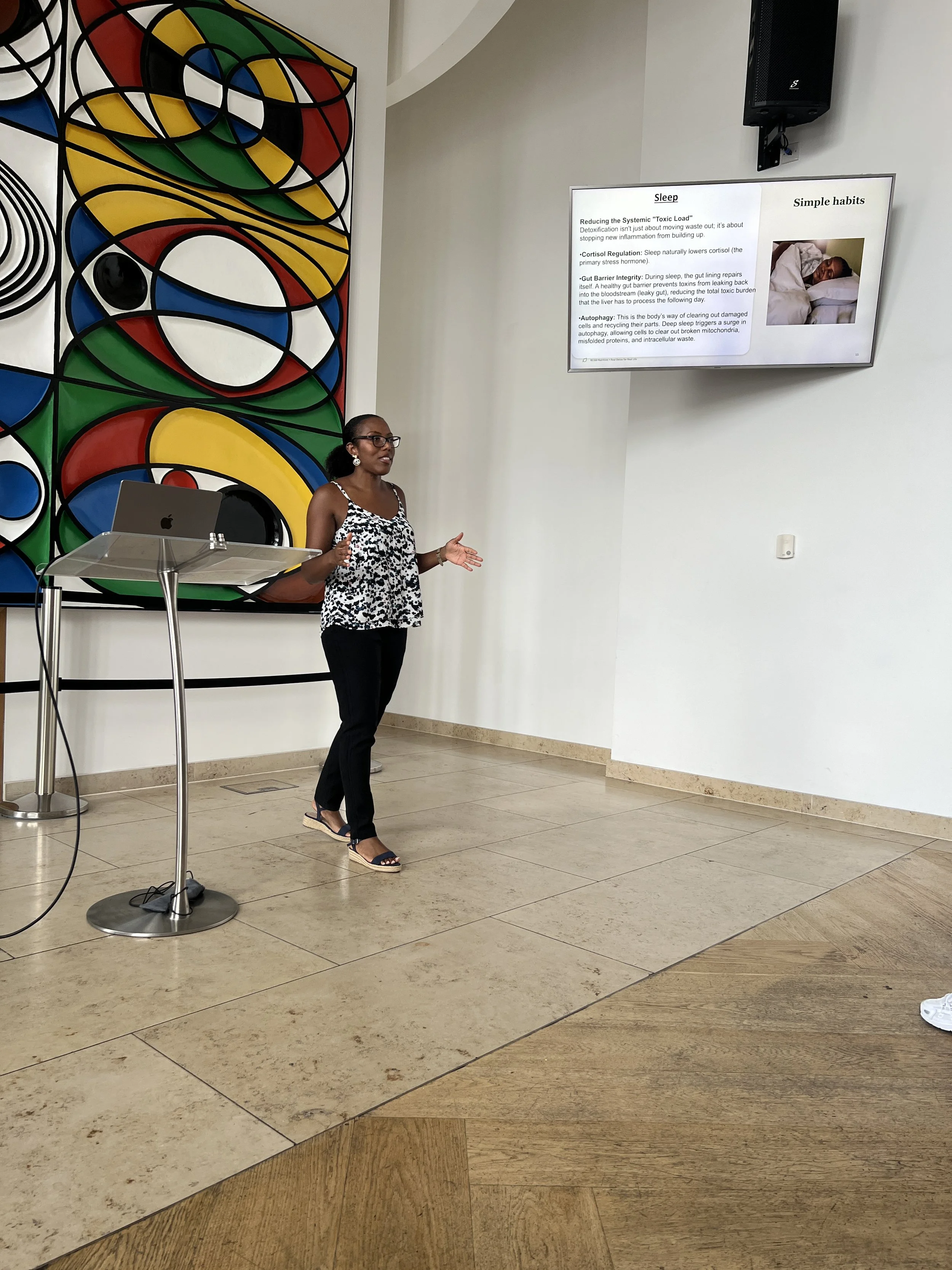 A woman giving a presentation in front of a large colorful abstract wall art and a screen with written text and an image of a sleeping person. She is wearing glasses, a black and white leopard print sleeveless top, black pants, and sandals. A laptop is on a stand nearby.