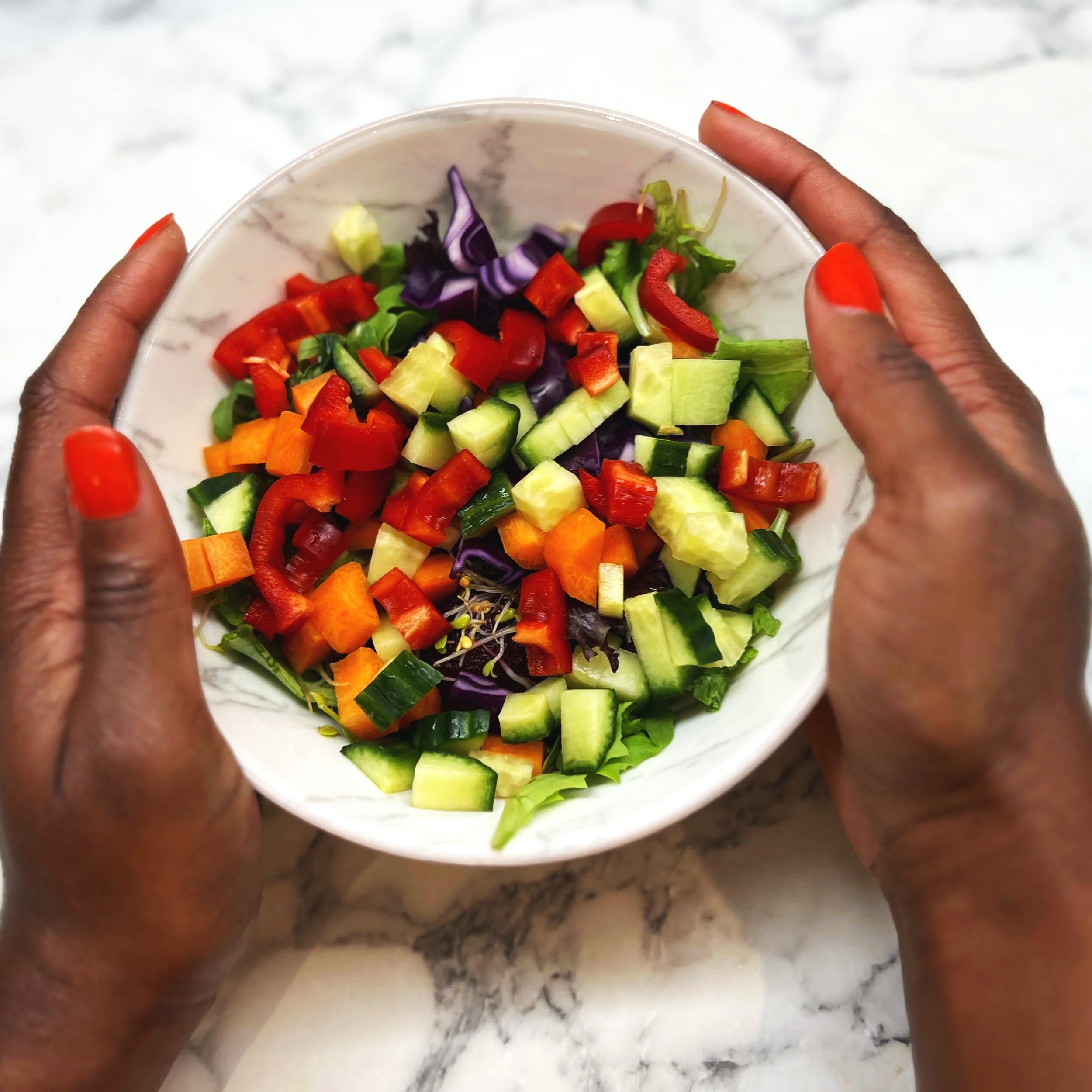 A person holding a white bowl with chopped vegetables, including red bell peppers, purple cabbage, cucumbers, and carrots, over a marble countertop.