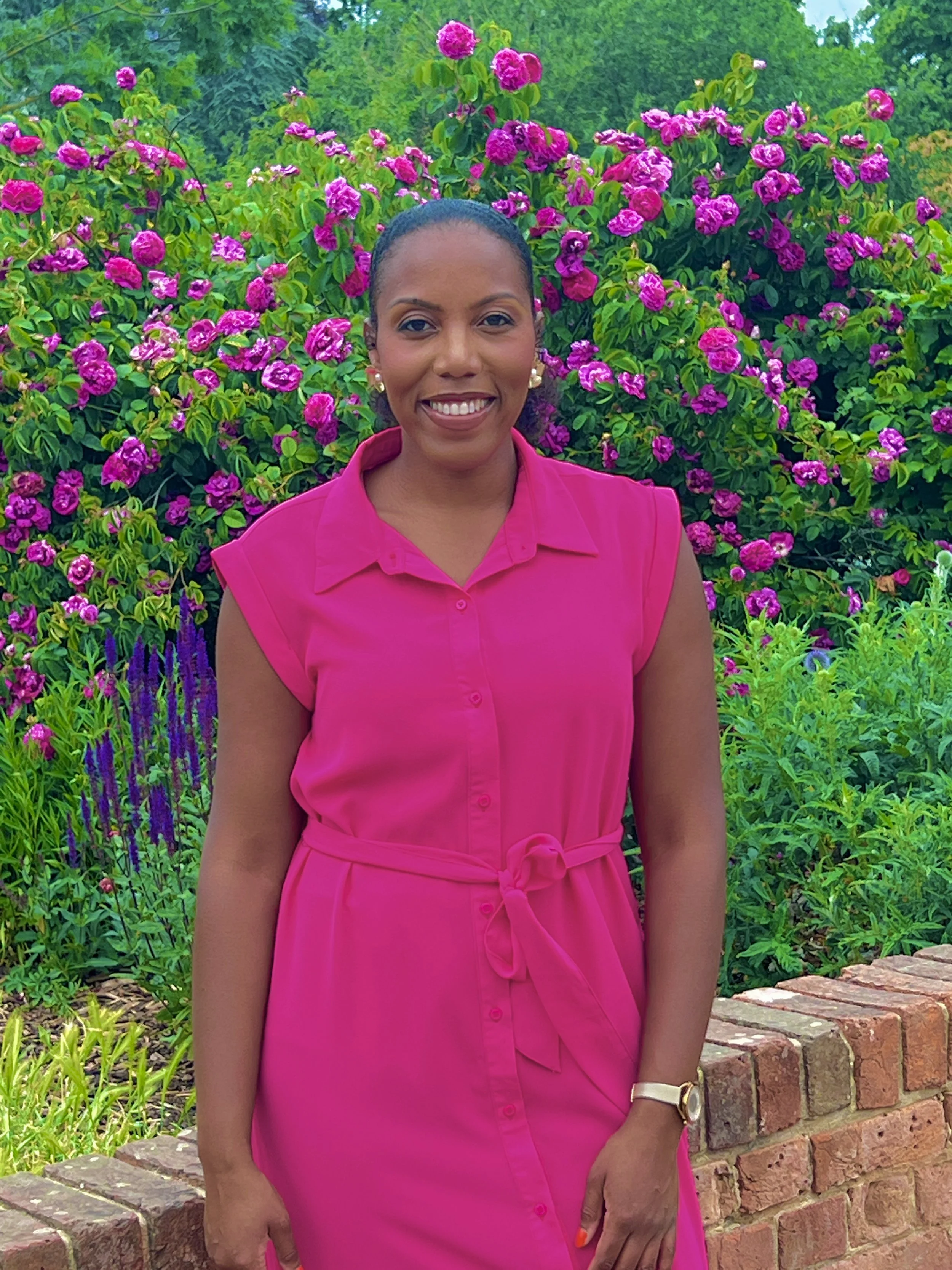 A woman wearing a pink dress and earrings standing in front of a flowering bush with pink flowers and green leaves.