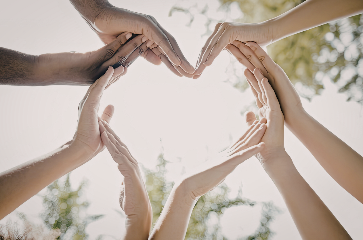 Multiple hands of different skin tones forming a heart shape against a bright sky and trees.