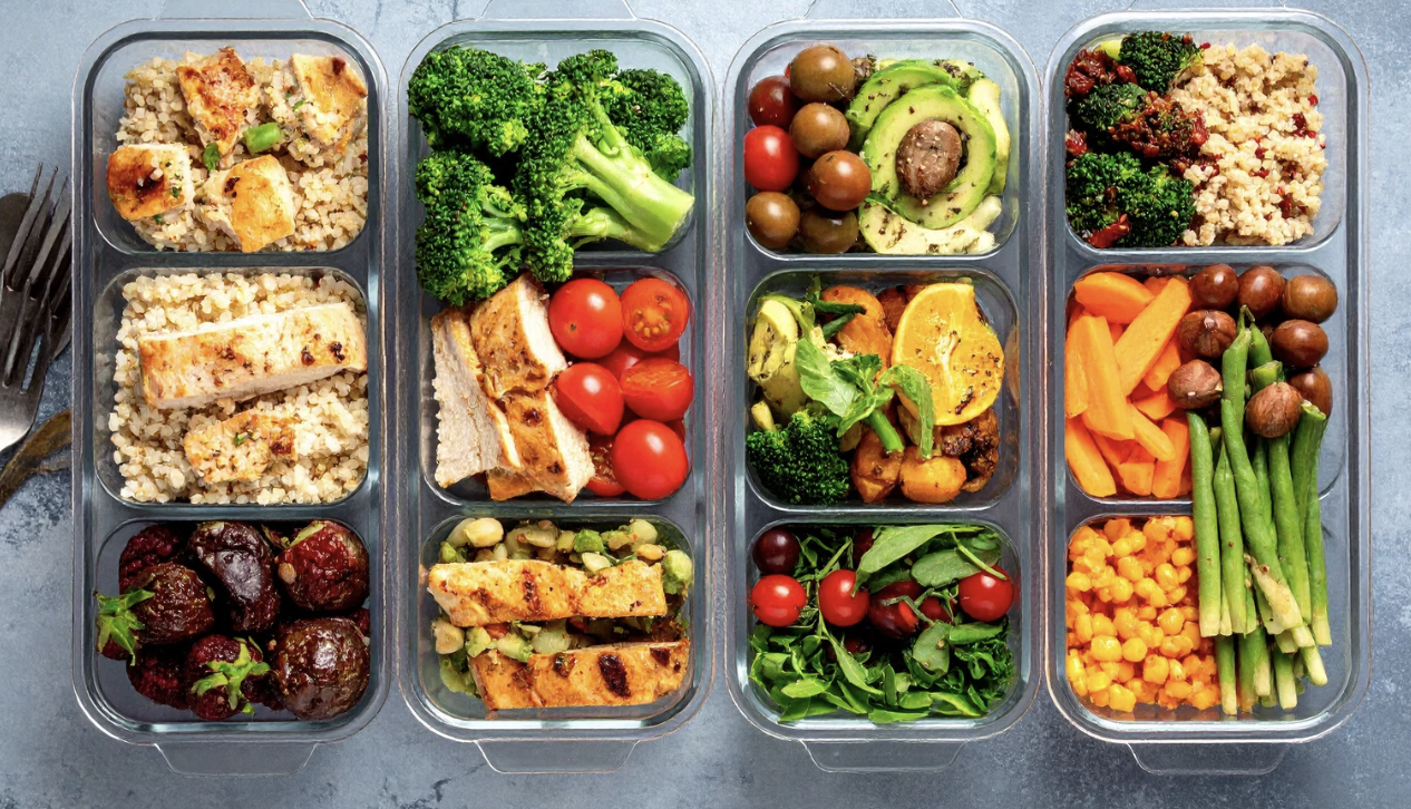 Four glass meal prep containers filled with various healthy foods including grains, vegetables, cherry tomatoes, grilled chicken, and fruit, arranged on a gray surface.
