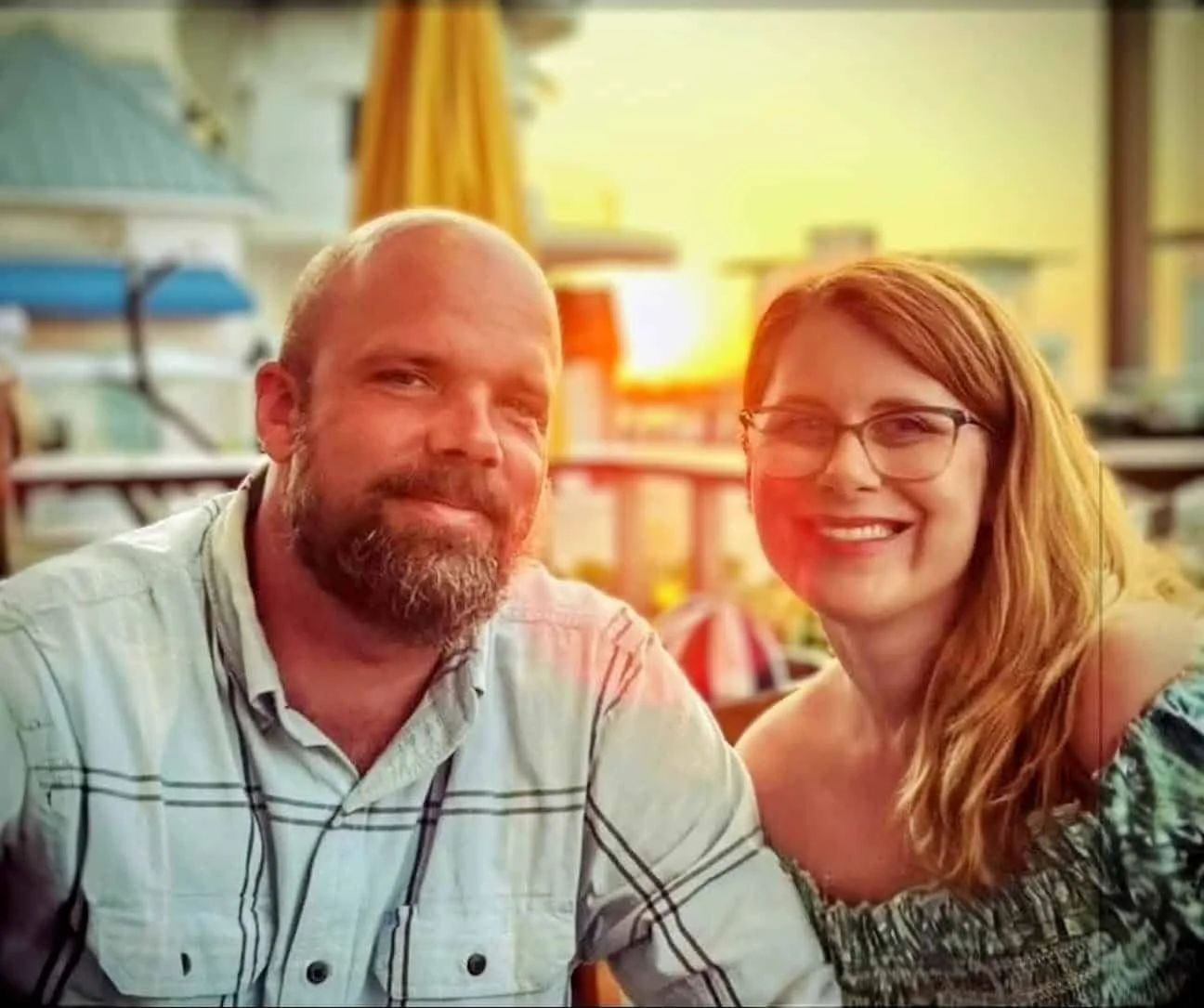 A man with a beard and a woman with glasses smiling at the camera indoors with warm lighting and colorful background decor.