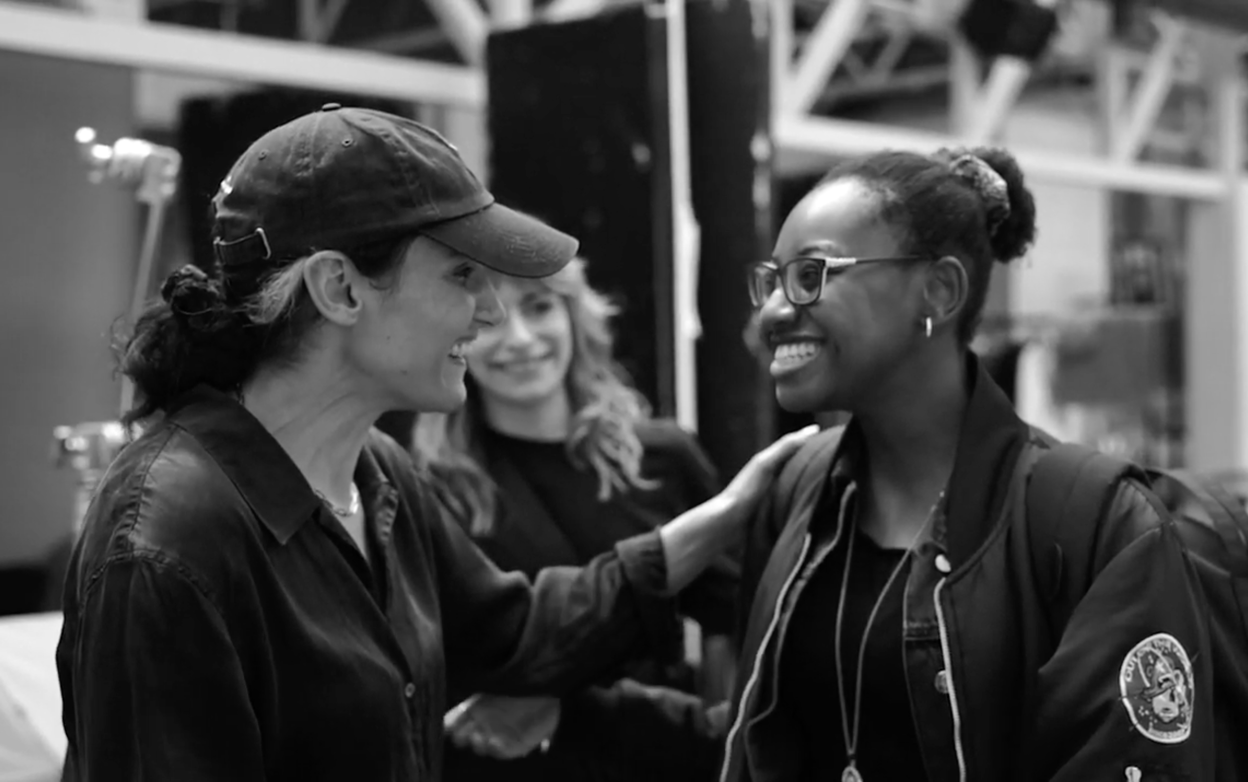 A black and white photo of two women smiling at each other, with one wearing glasses and the other wearing a cap.