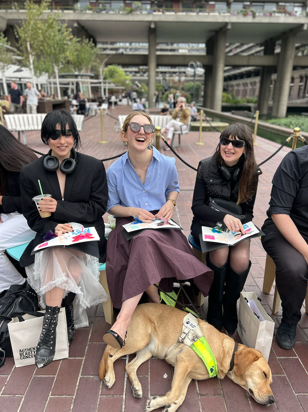 Three laughing guests with tactile booklets sit in the front row, a guide dog asleep at their feet.