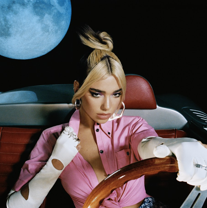 A woman with blonde hair styled in a top bun and wearing a pink shirt, sitting in a car with a moon visible through the back window.