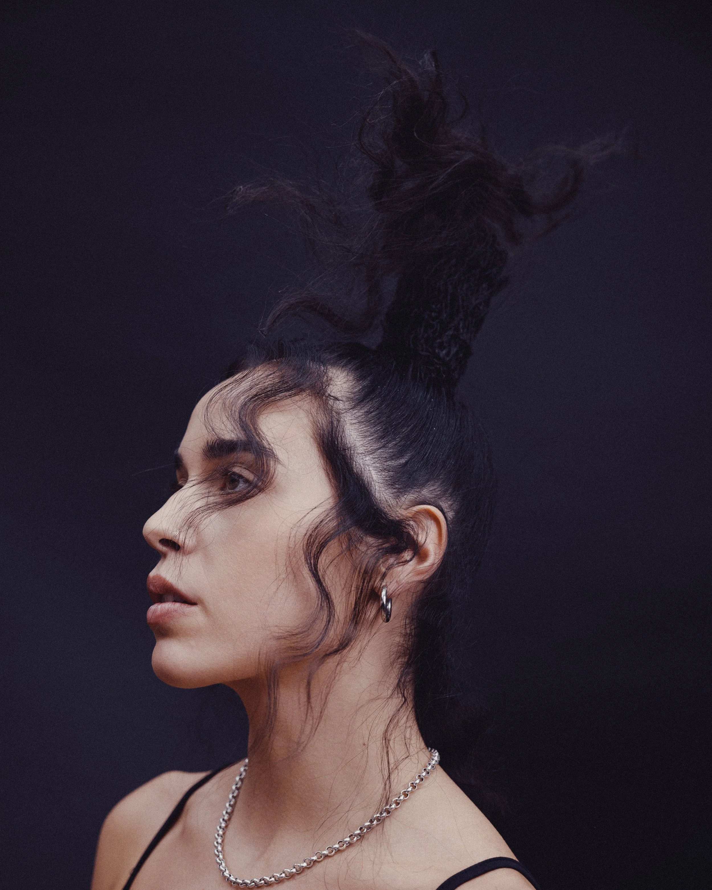 A woman with dark, curly hair styled high and upward, wearing a black spaghetti strap top, hoop earrings, and layered silver necklaces, against a dark background.