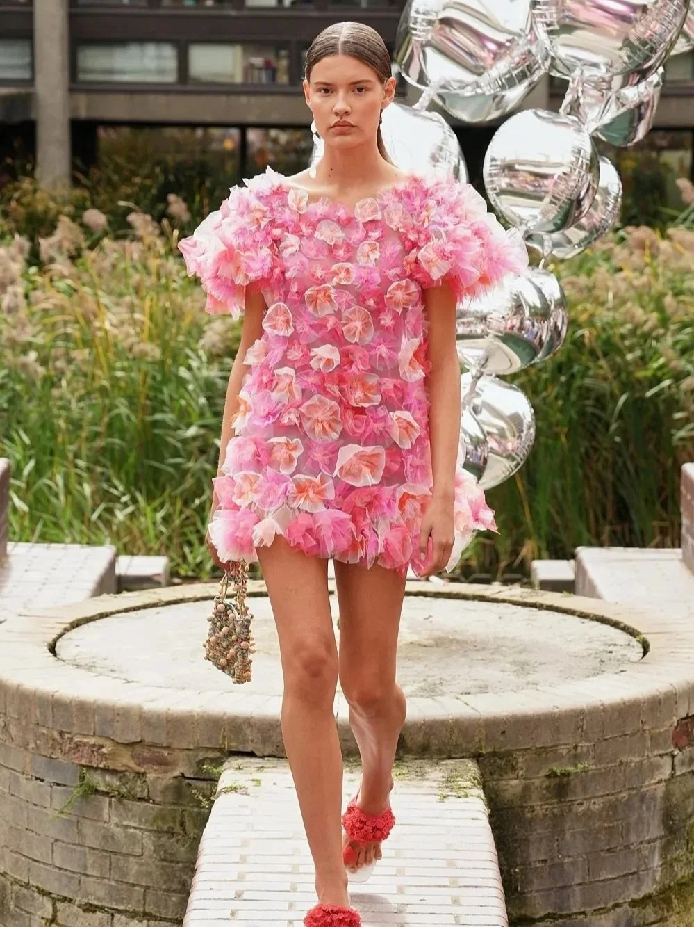 A model wearing a pink floral dress and matching sandals walks along an outdoor runway surrounded by greenery and silver balloon sculptures.