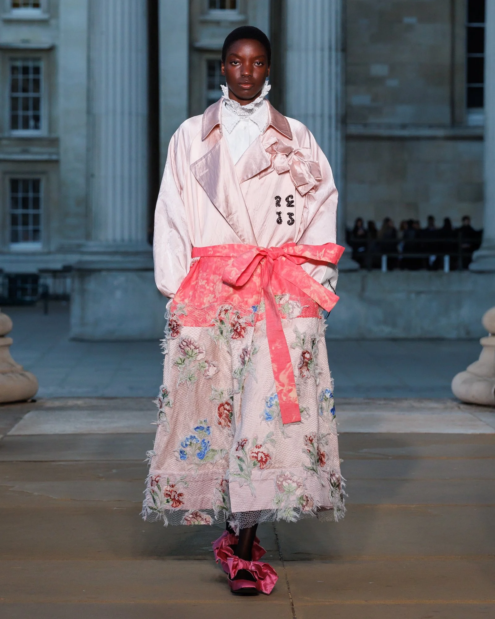 A model walks on a runway wearing a pastel pink and floral outfit with large pink ruffled slippers, set against a backdrop of classical building columns and windows.