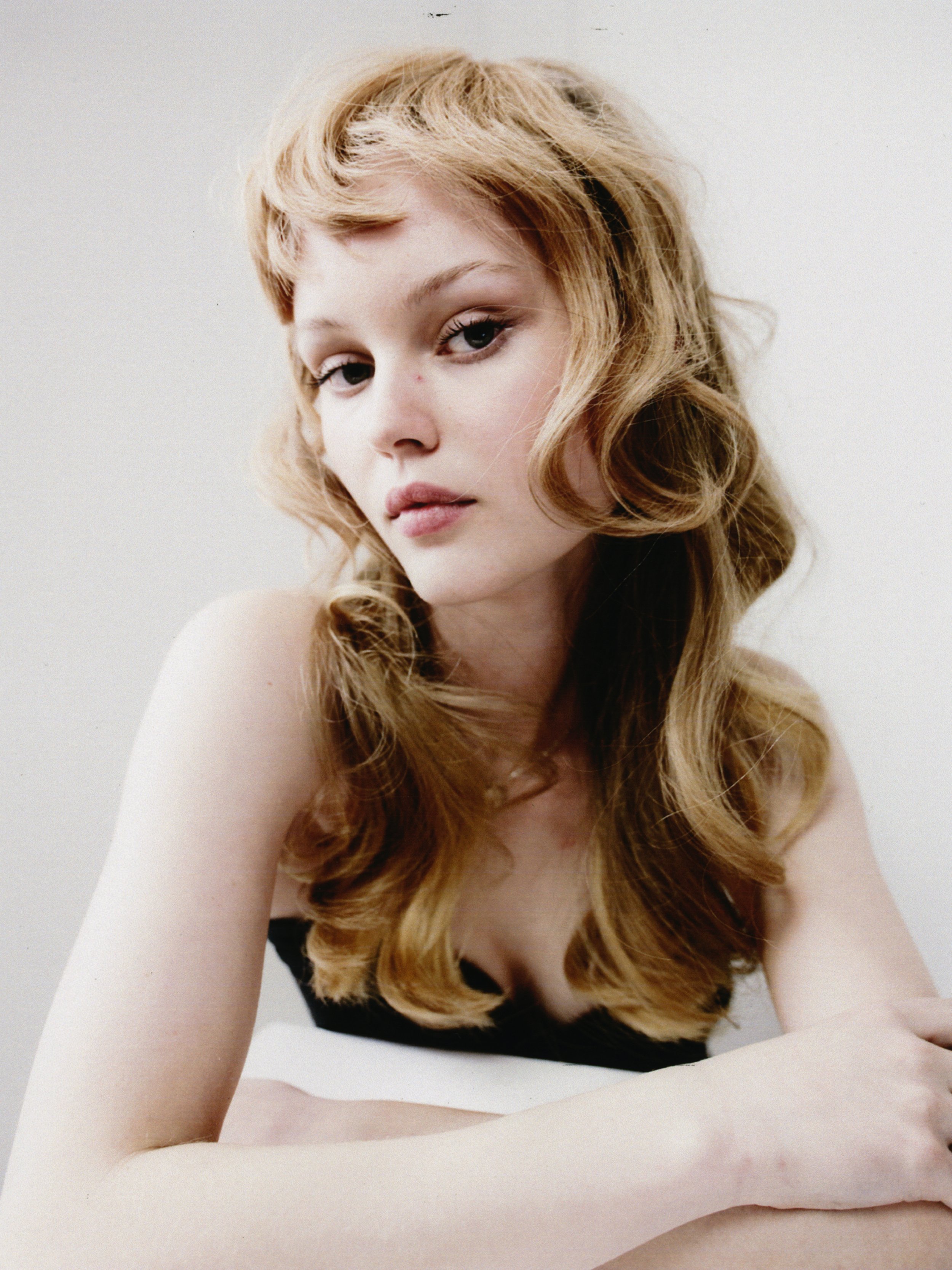 A young woman with long, wavy light brown hair and fair skin, looking towards the camera with a neutral expression, against a plain white background.