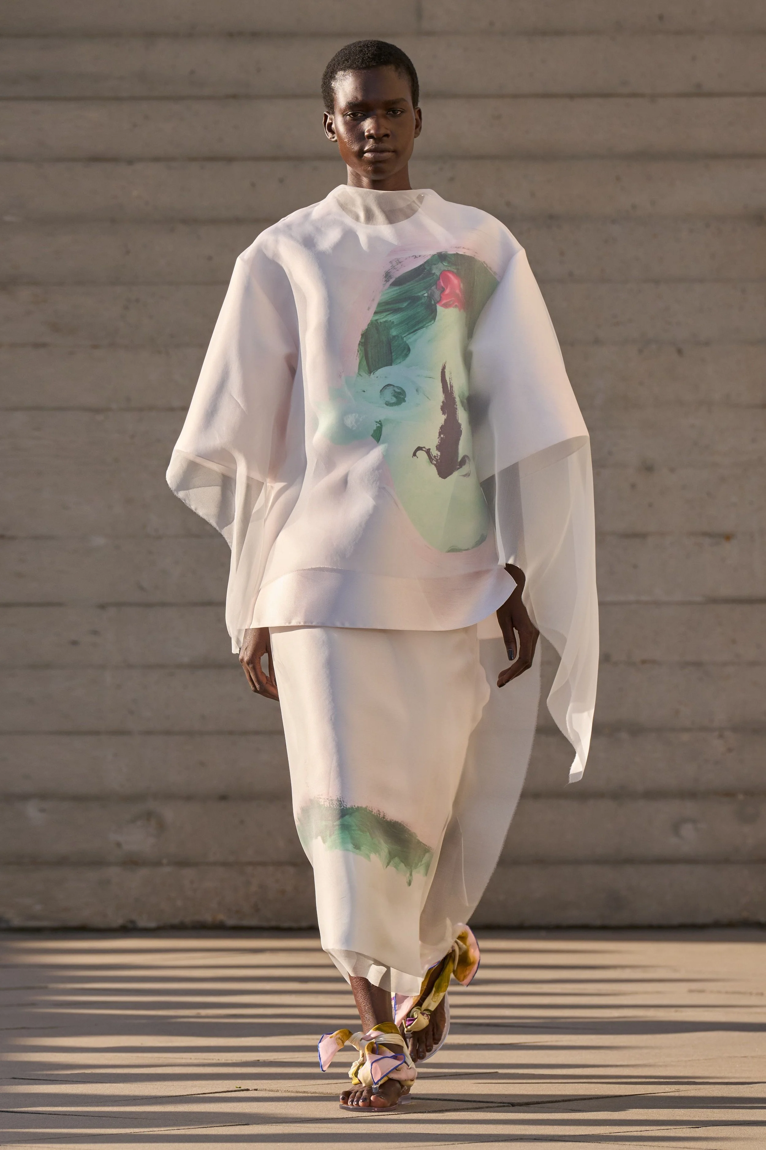 A model in a sheer white top and skirt with a painterly green and pink print walks in low sunlight.