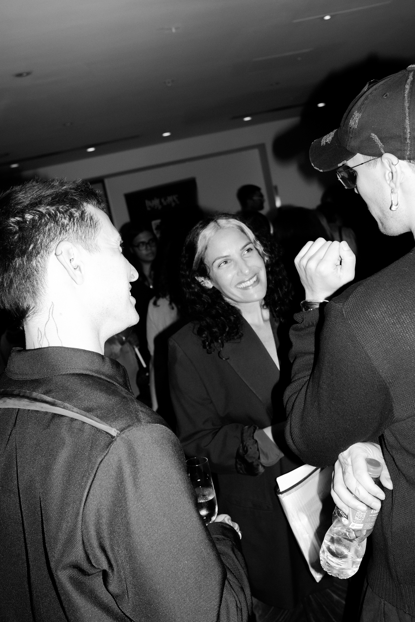 A group of people gathered at an indoor event, with a woman smiling while talking with a man in the foreground.