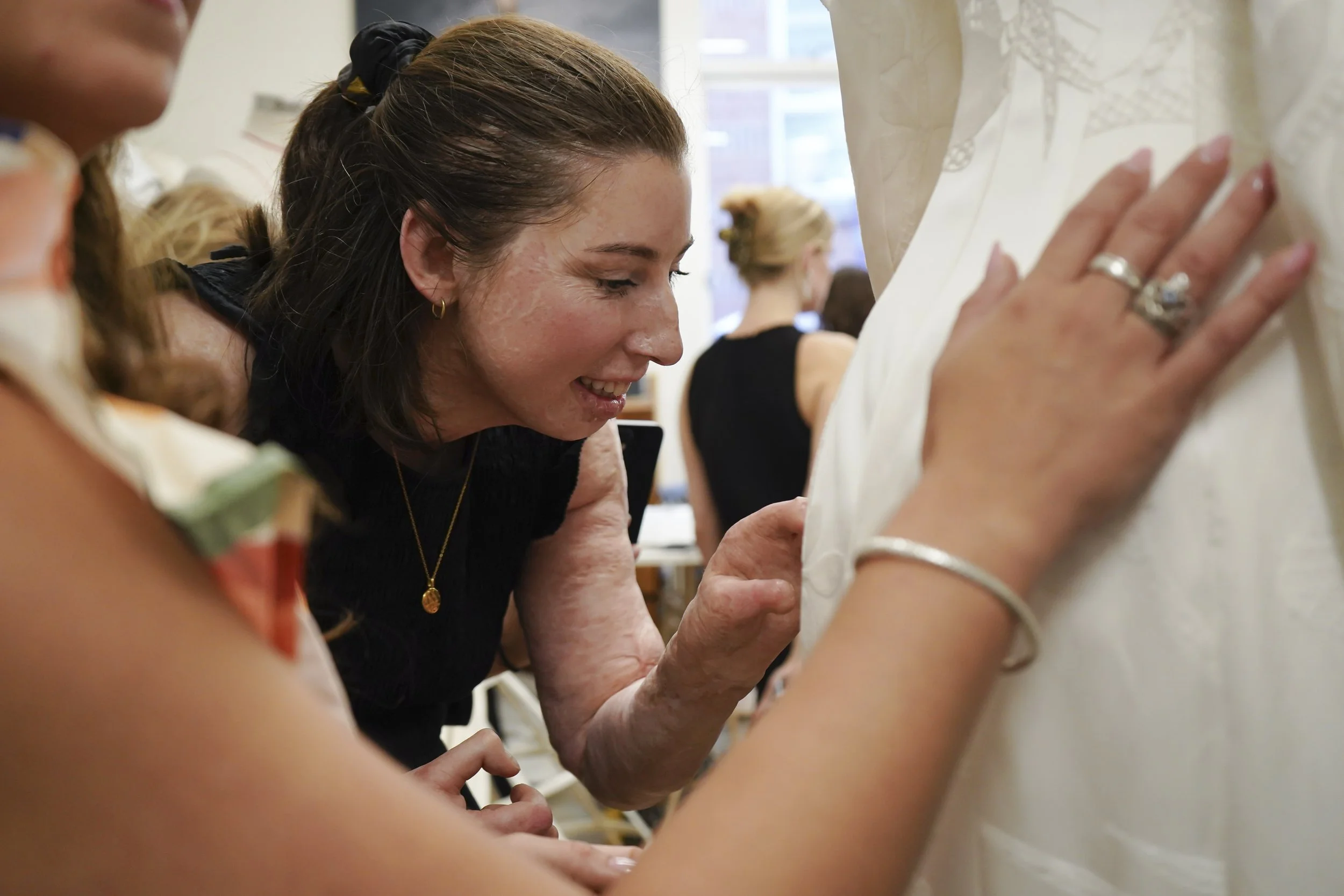 A smiling woman leans in to feel the embroidered detail on a white gown. Another person's arm can be seen in the foreground, doing the same.
