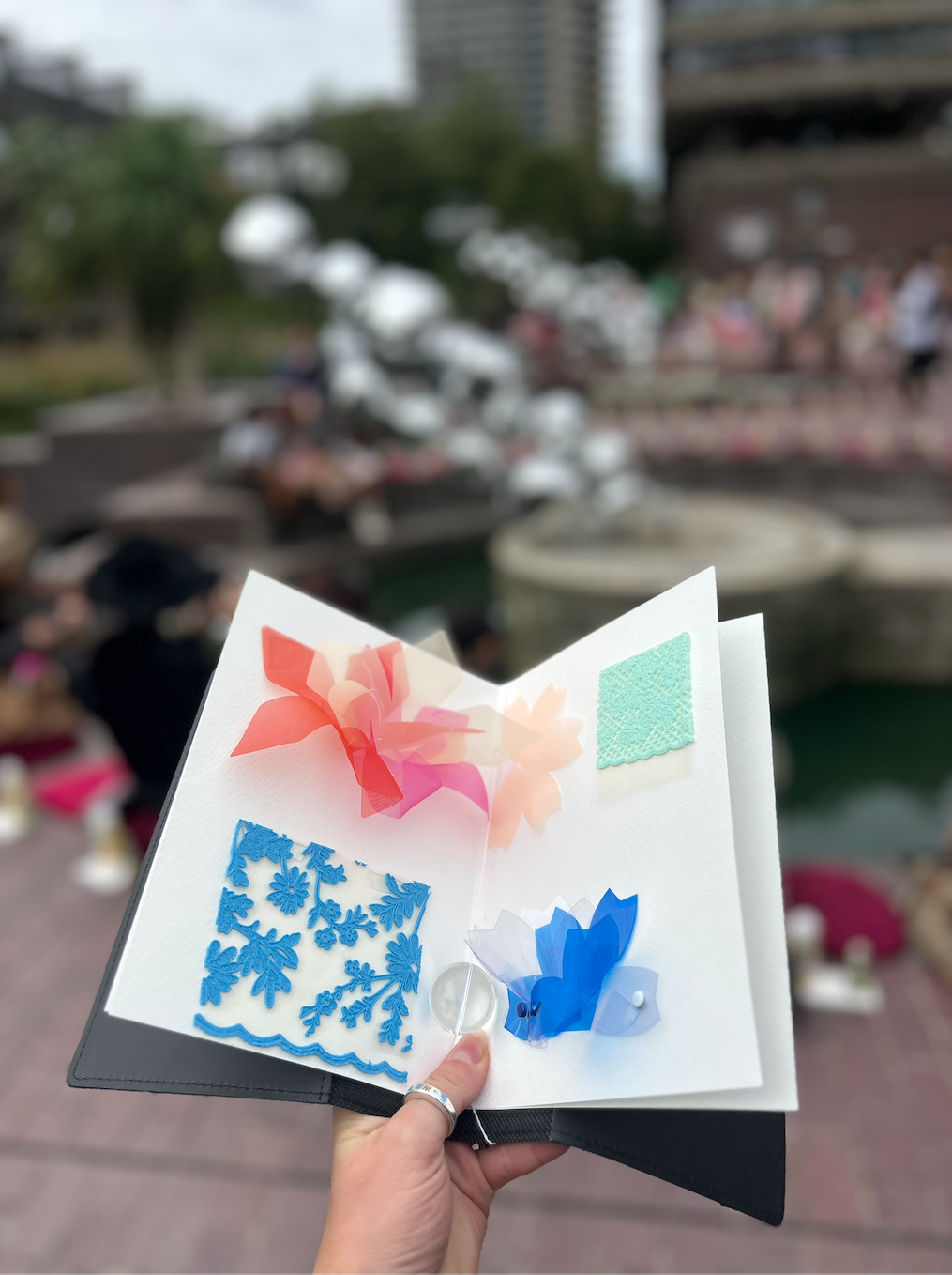 A hand holds open a tactile booklet with petal, lace and bead swatches at an outdoor show.