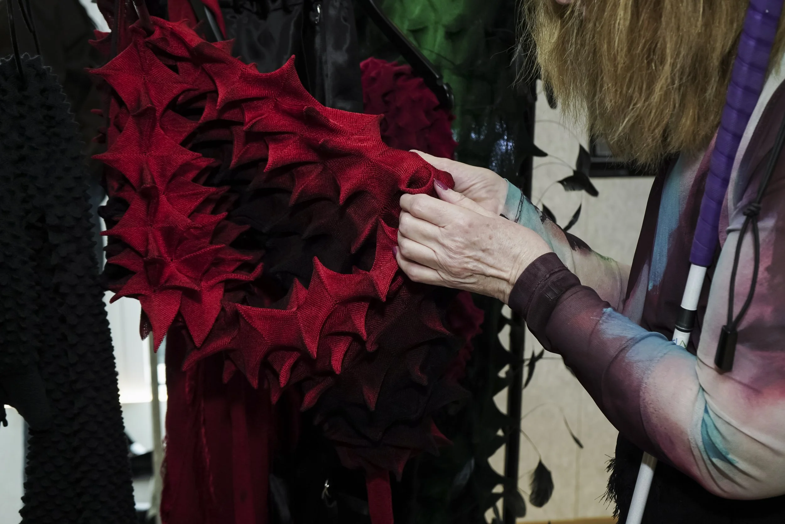 A close-up of a person's hands examining a red spiky knitted dress, hung on a rail beside a similarly spiky black garment