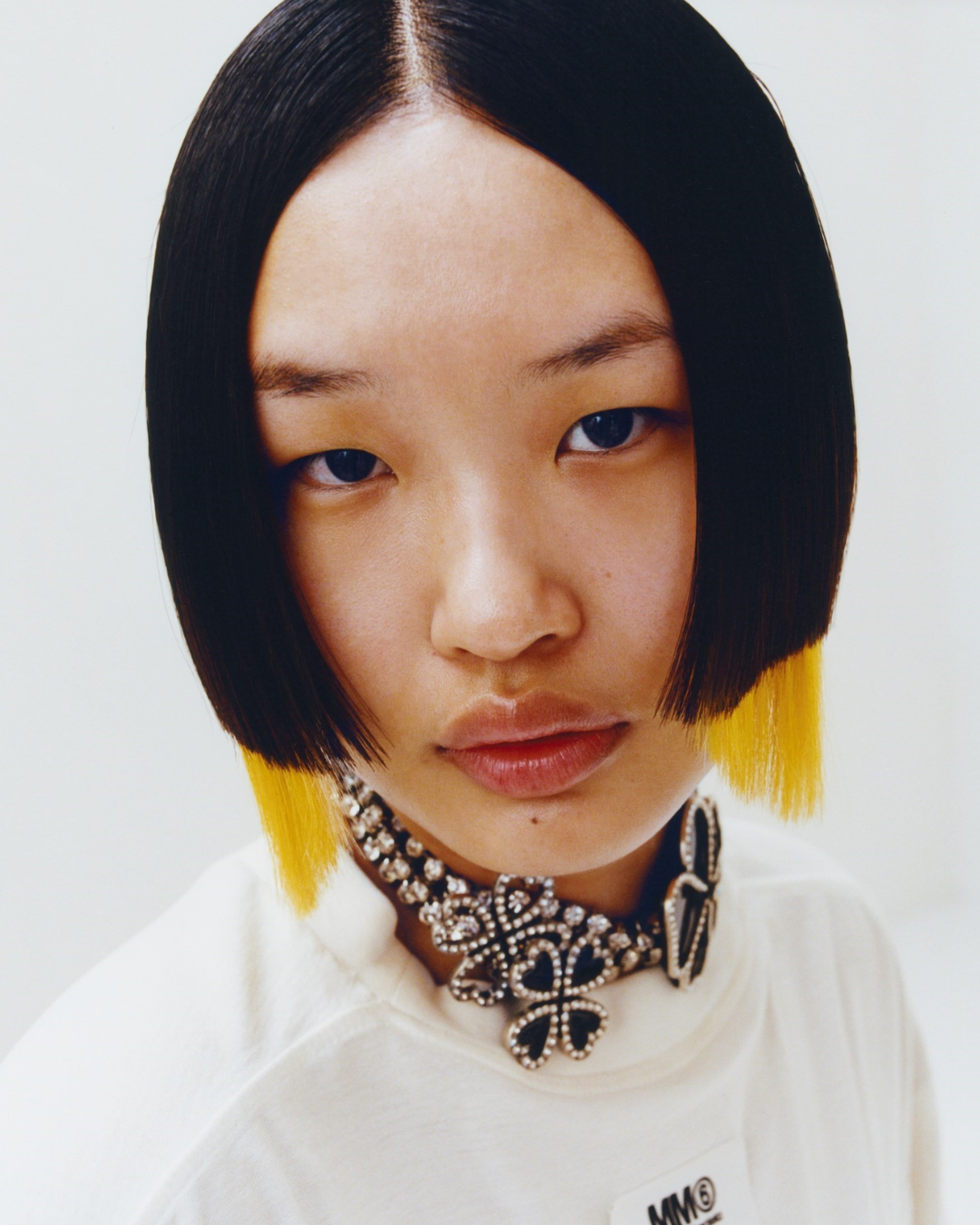 Close-up of a young woman with straight black hair, wearing a white top including a large statement necklace with black and silver embellishments and yellow dye at the tips of her hair. She has a neutral expression.