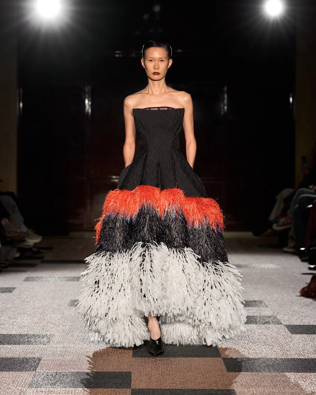 Model walking down runway in a strapless black, red, black, and white feathered gown at a fashion show.