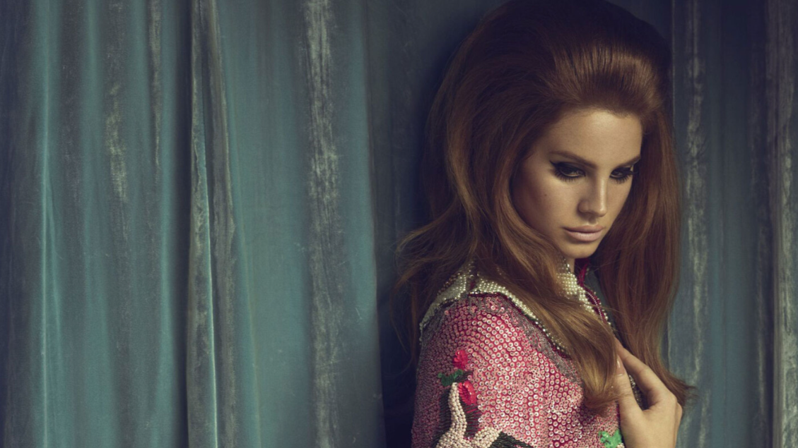 A picture of Lana Del Ray with wavy brown hair, wearing a pink and silver beaded top with floral embroidery, standing in front of teal curtains, looking downward with a contemplative expression.