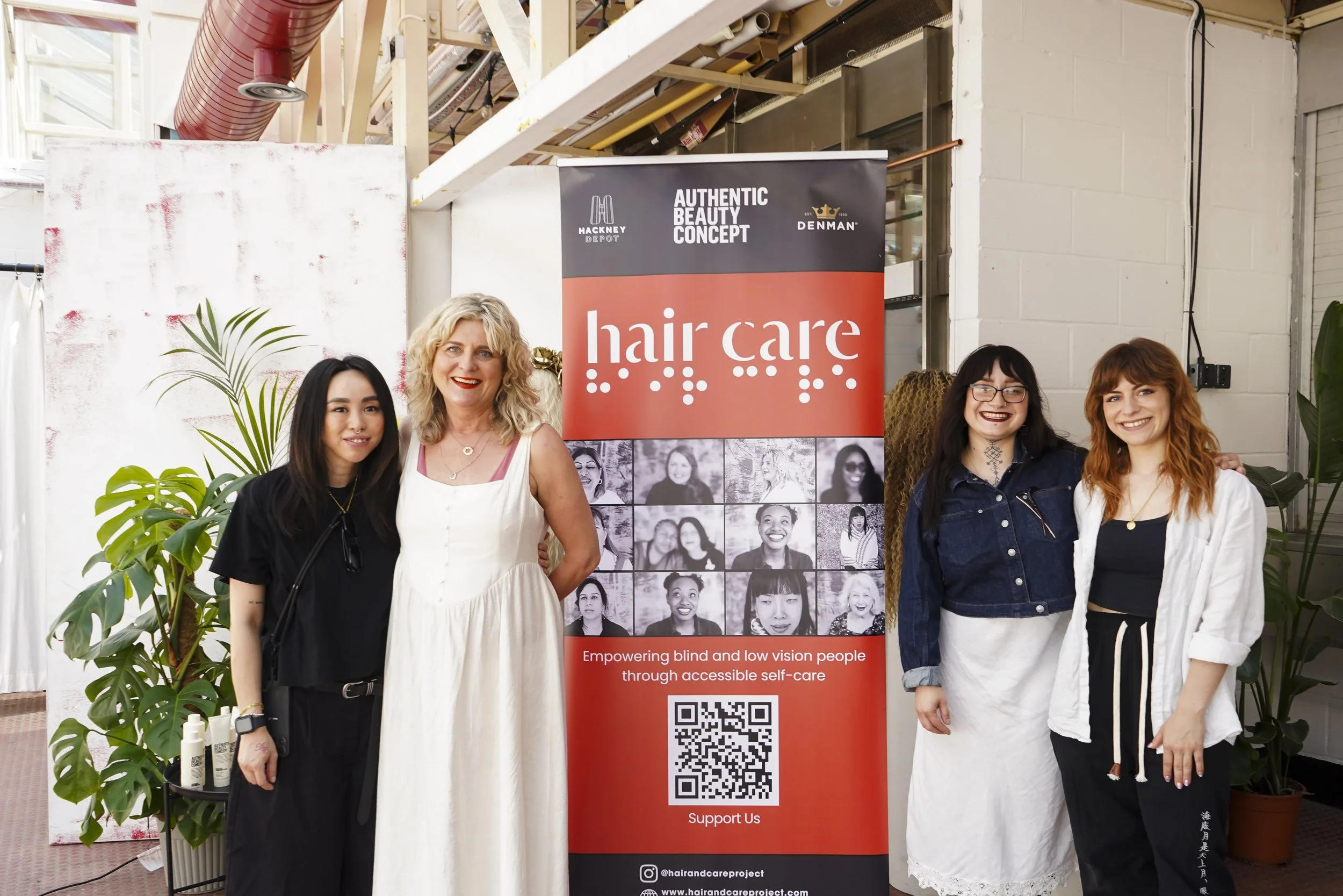 Four women smile beside a banner about accessible self-care at the Hair & Care workshop, in a bright industrial space.