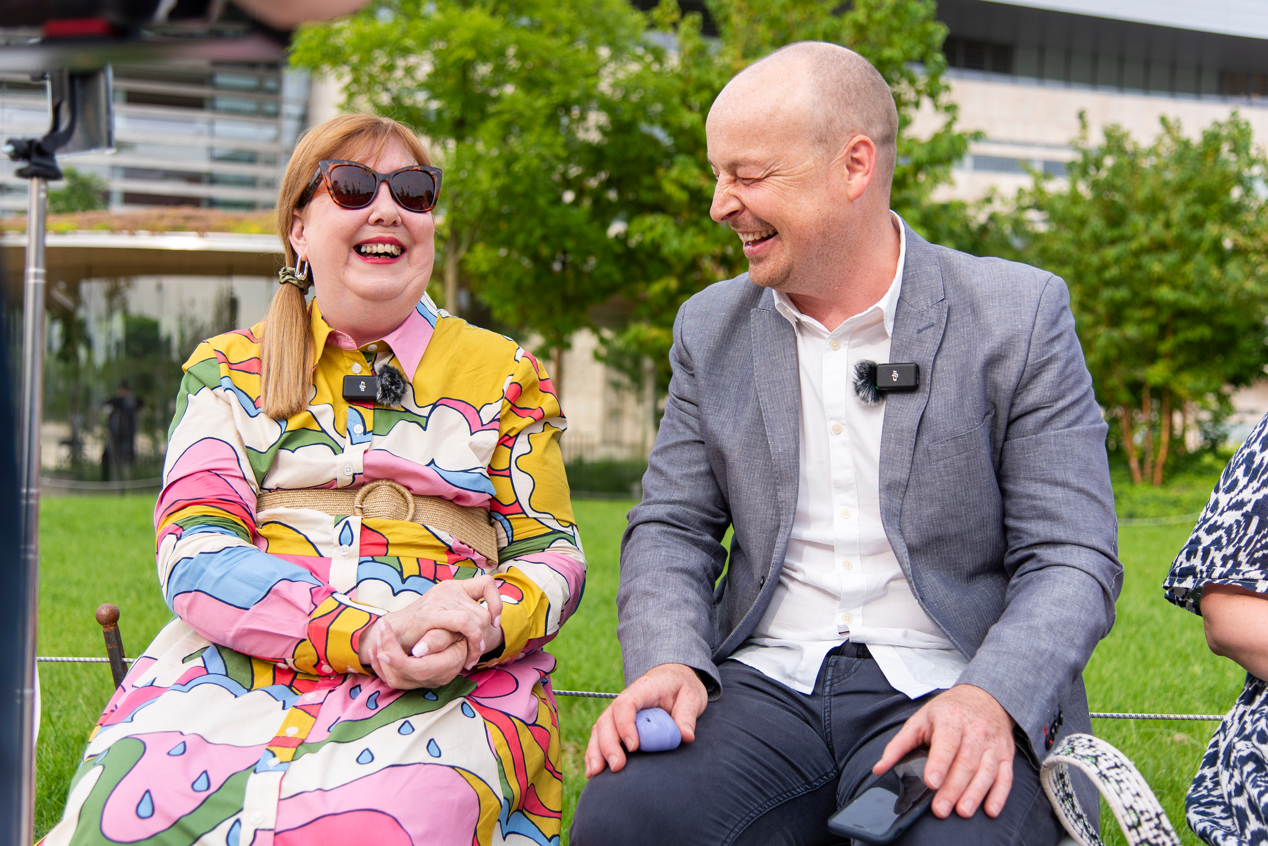Two guests at a fashion show are sitting and laughing together outdoors in a park with green trees and buildings in the background.