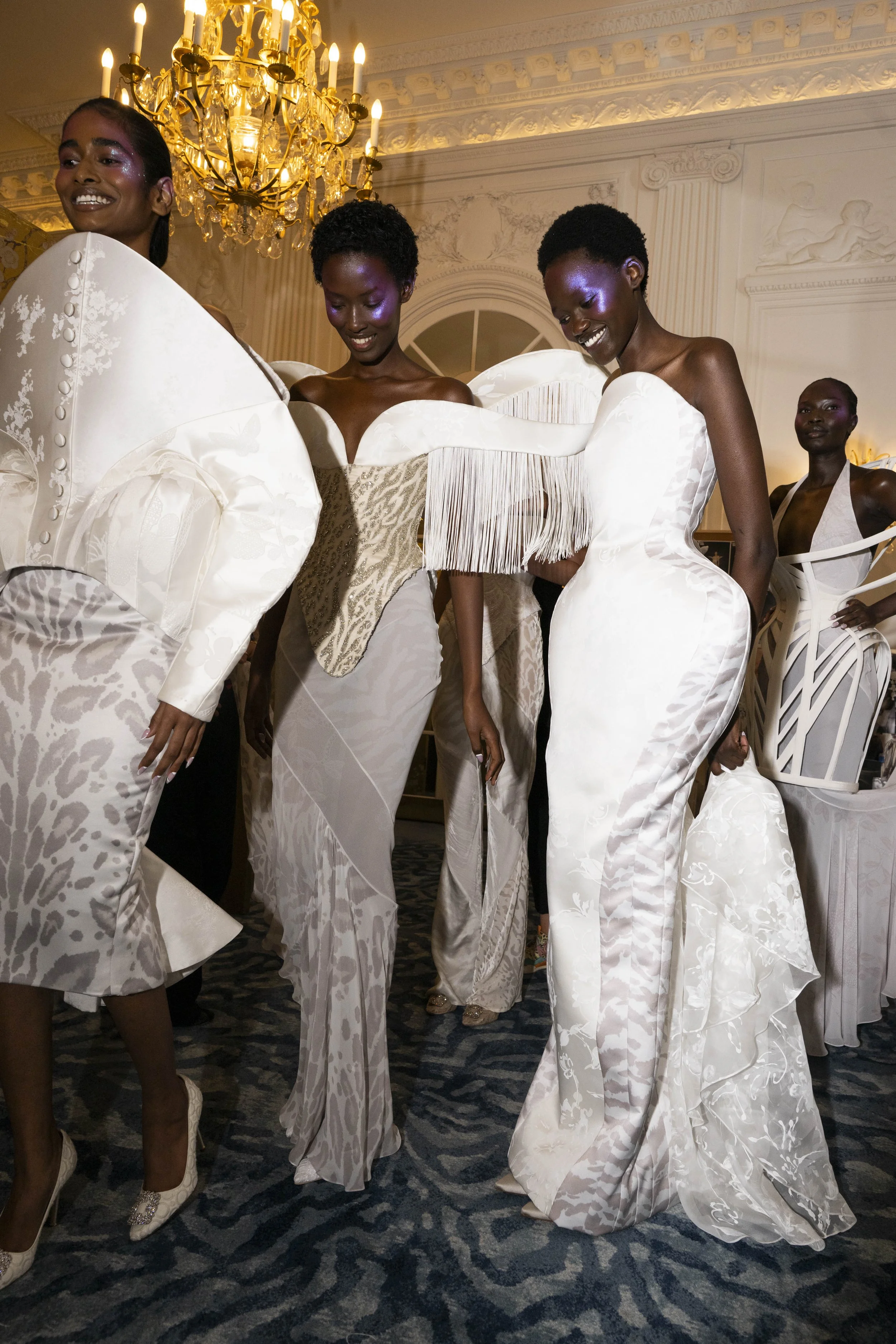 Smiling models in white sculptural gowns gather backstage beneath a chandelier.