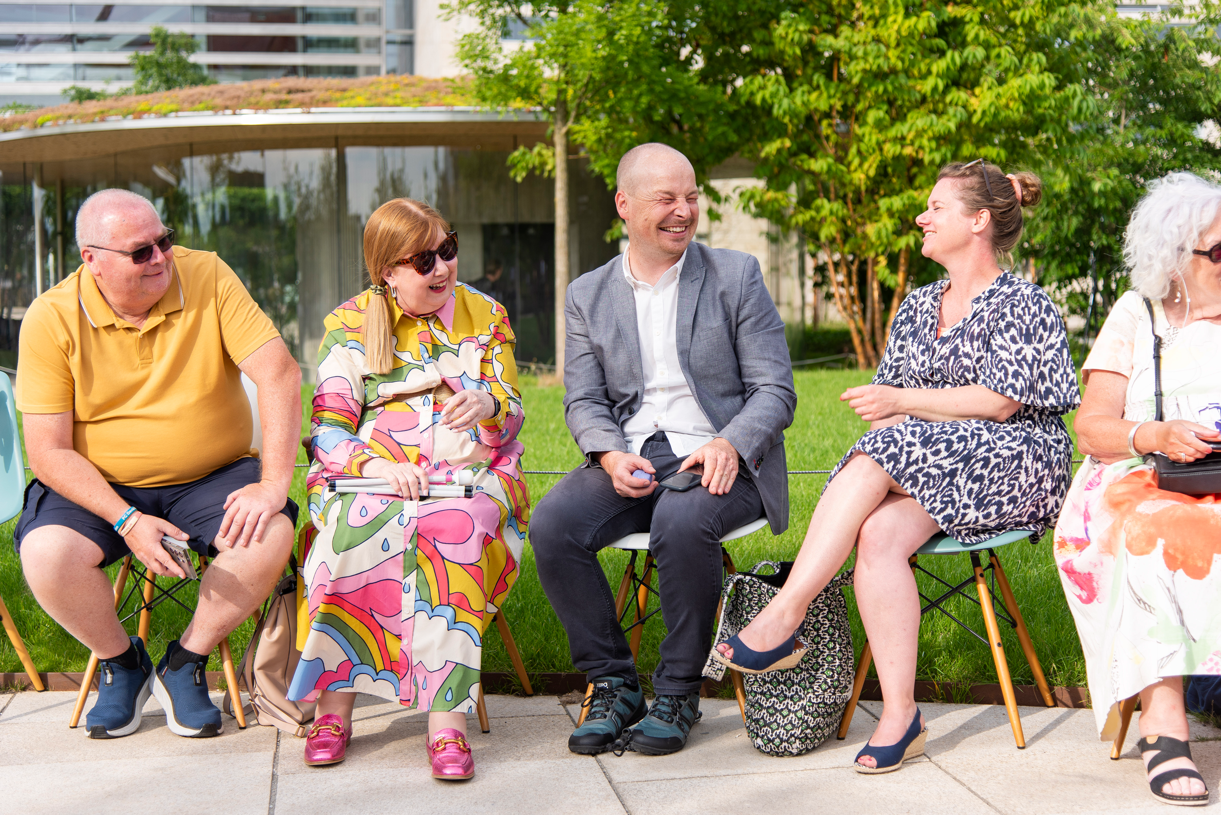 Group of five guests at a fashion show, seated outdoors on chairs, smiling and chatting, with a building and green trees in the background, and the runway in the foreground.