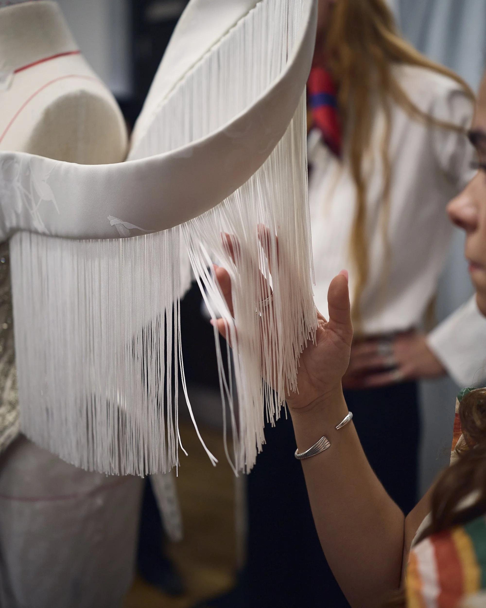 A hand gently feels the long white fringe on a gown displayed on a mannequin.