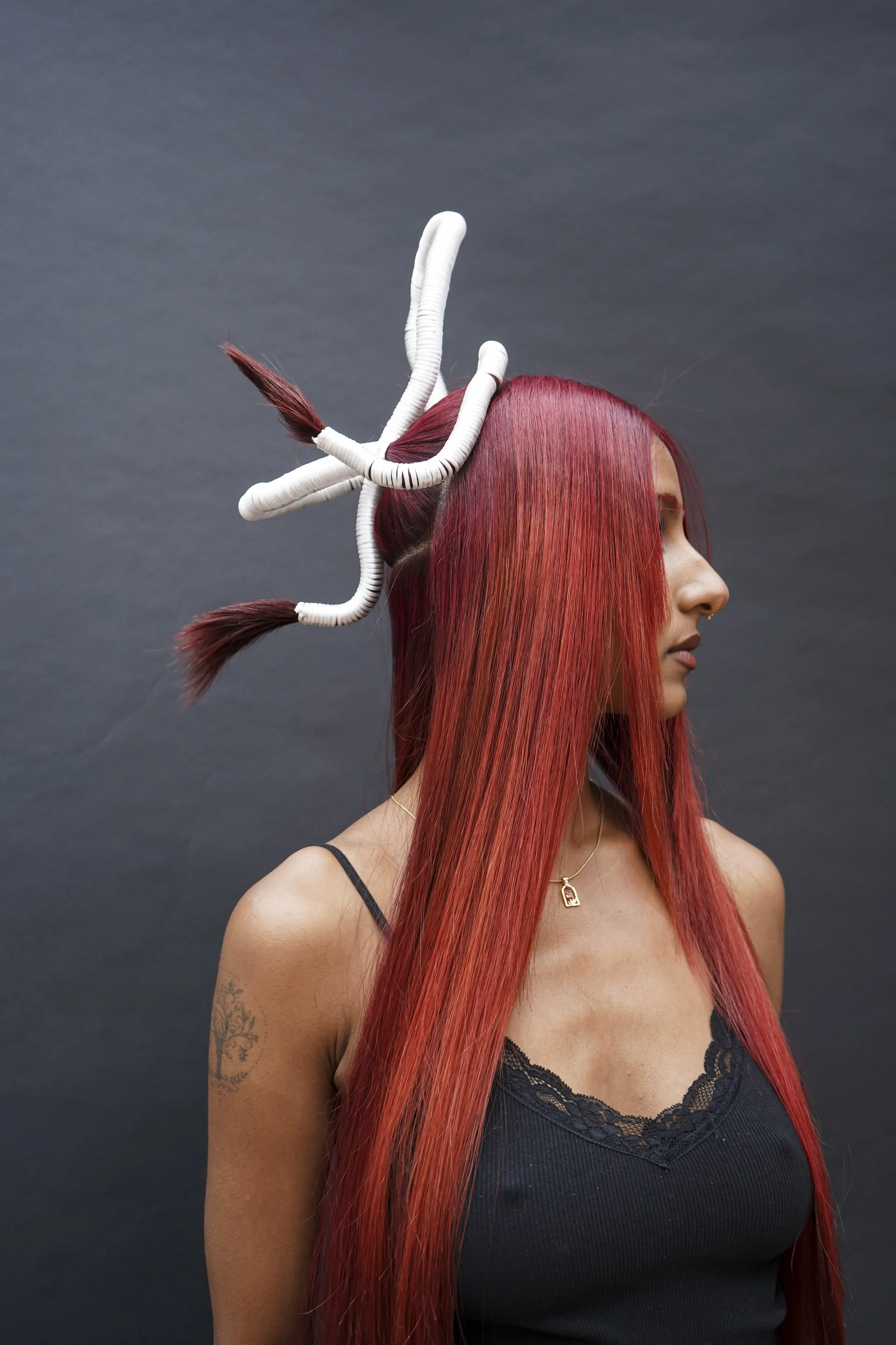 A woman with long, straight, vibrant red hair with a section styled into white twisting shapes, against a dark grey background.