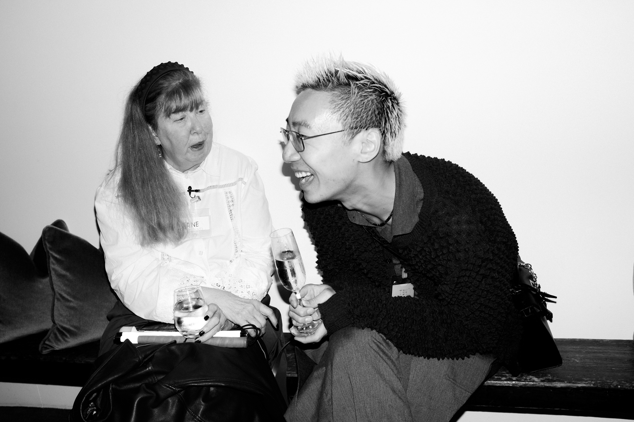 A black and white photo of a middle-aged woman and a young man laughing together, seated on a black bench against a white wall.