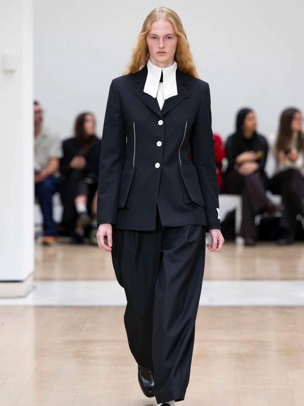 Model walking on runway wearing a black top, checkered coat, and sheer white pleated pants.