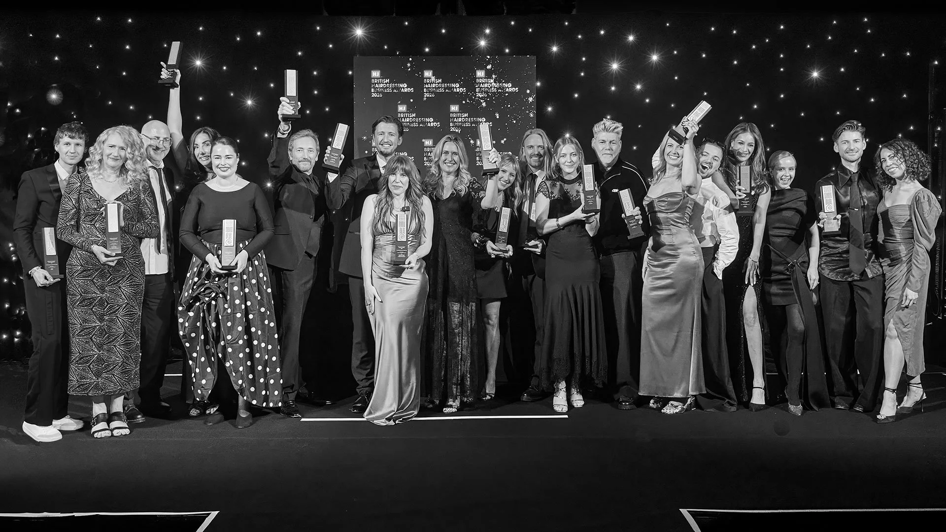 The Hair & Care team on stage at an awards ceremony, holding trophies, smiling, dressed in formal attire, with a backdrop displaying text related to the British Hairdressing Business Awards.